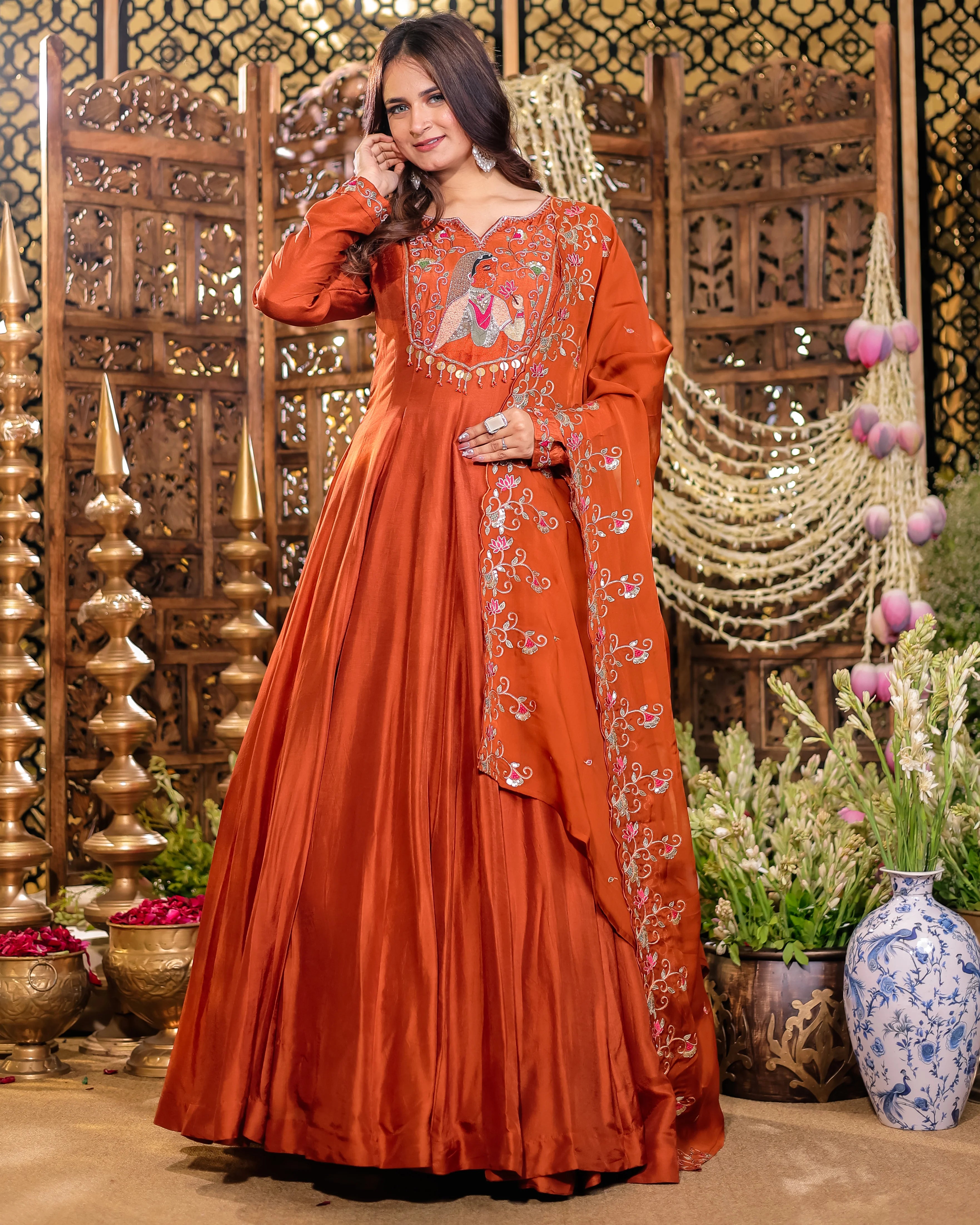 Woman in an orange embroidered traditional outfit standing in a decorative setting with plants and vases.
