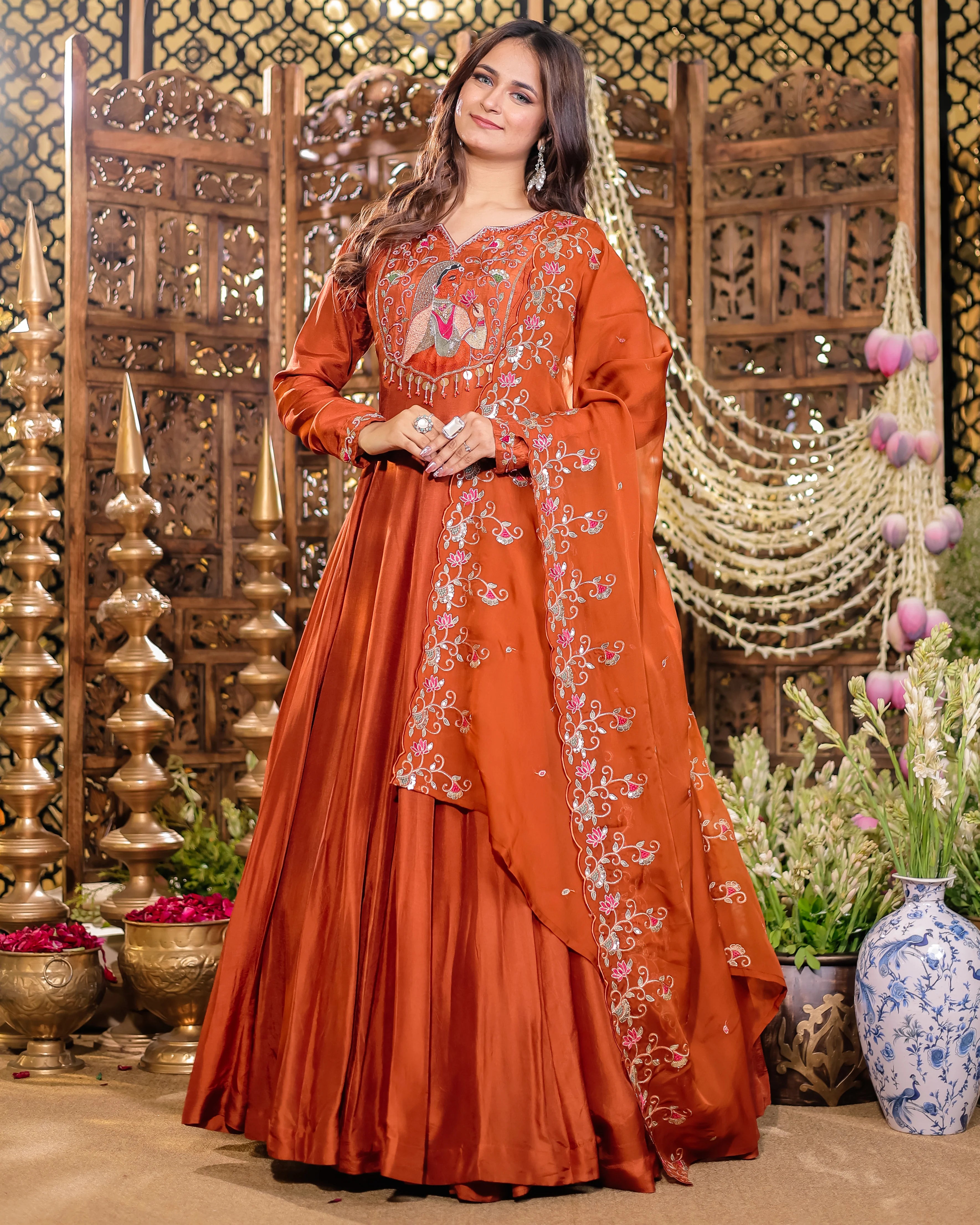 Woman in an orange embroidered traditional outfit standing in a decorative setting with wooden carvings and flowers.
