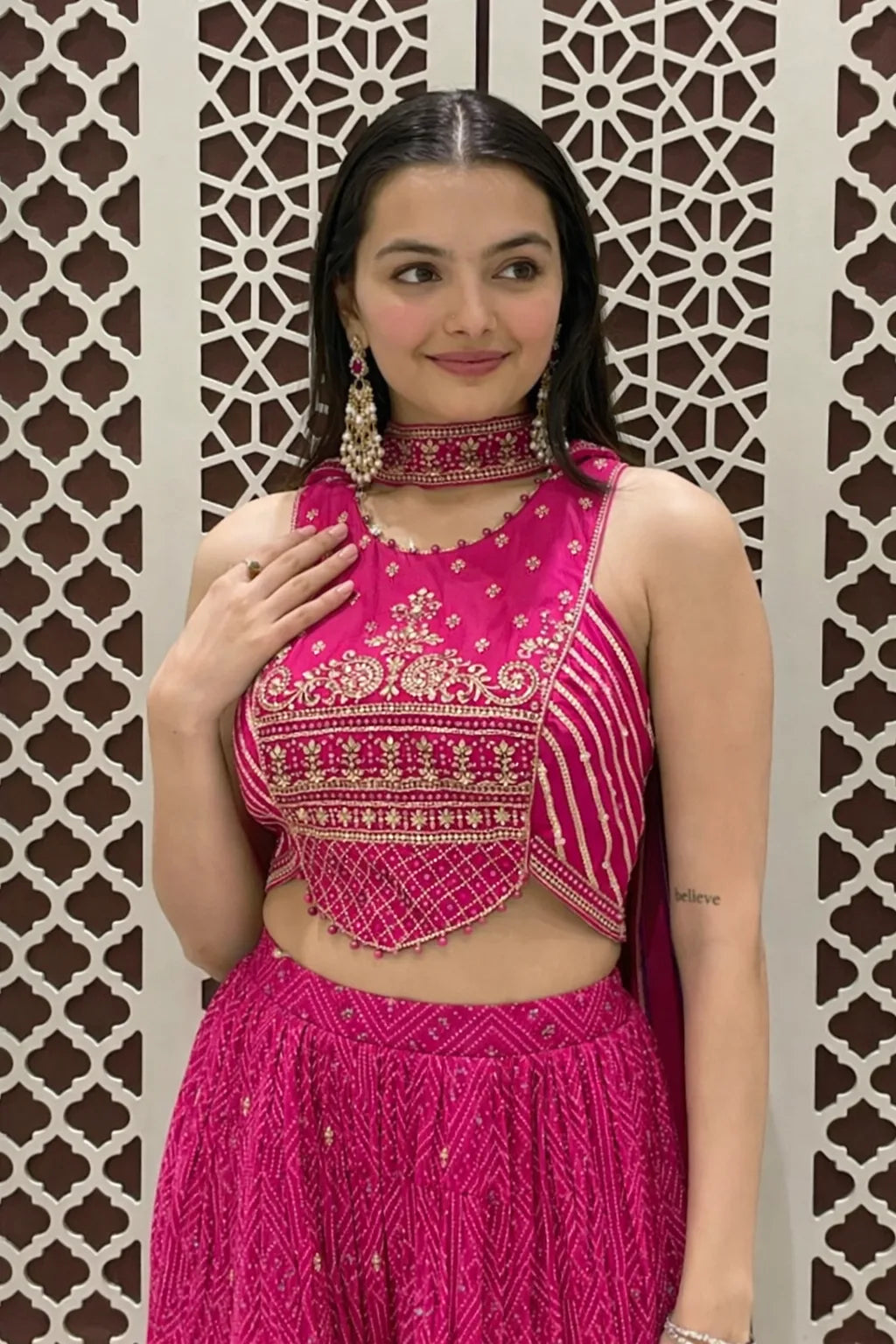 Woman wearing a bright pink traditional outfit with intricate designs against a decorative wall.