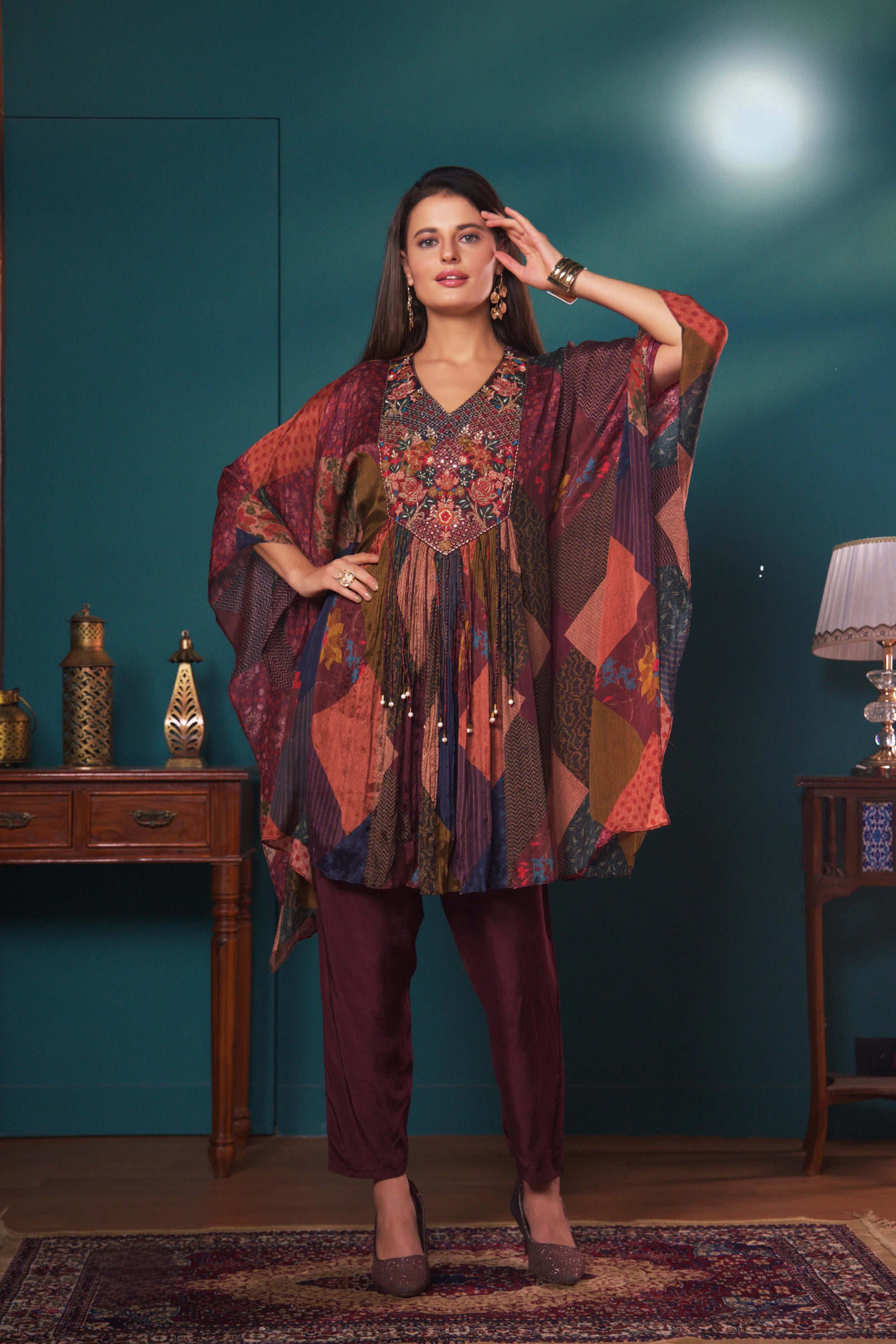 Woman in a patterned outfit standing in a room with wooden furniture and a lamp.