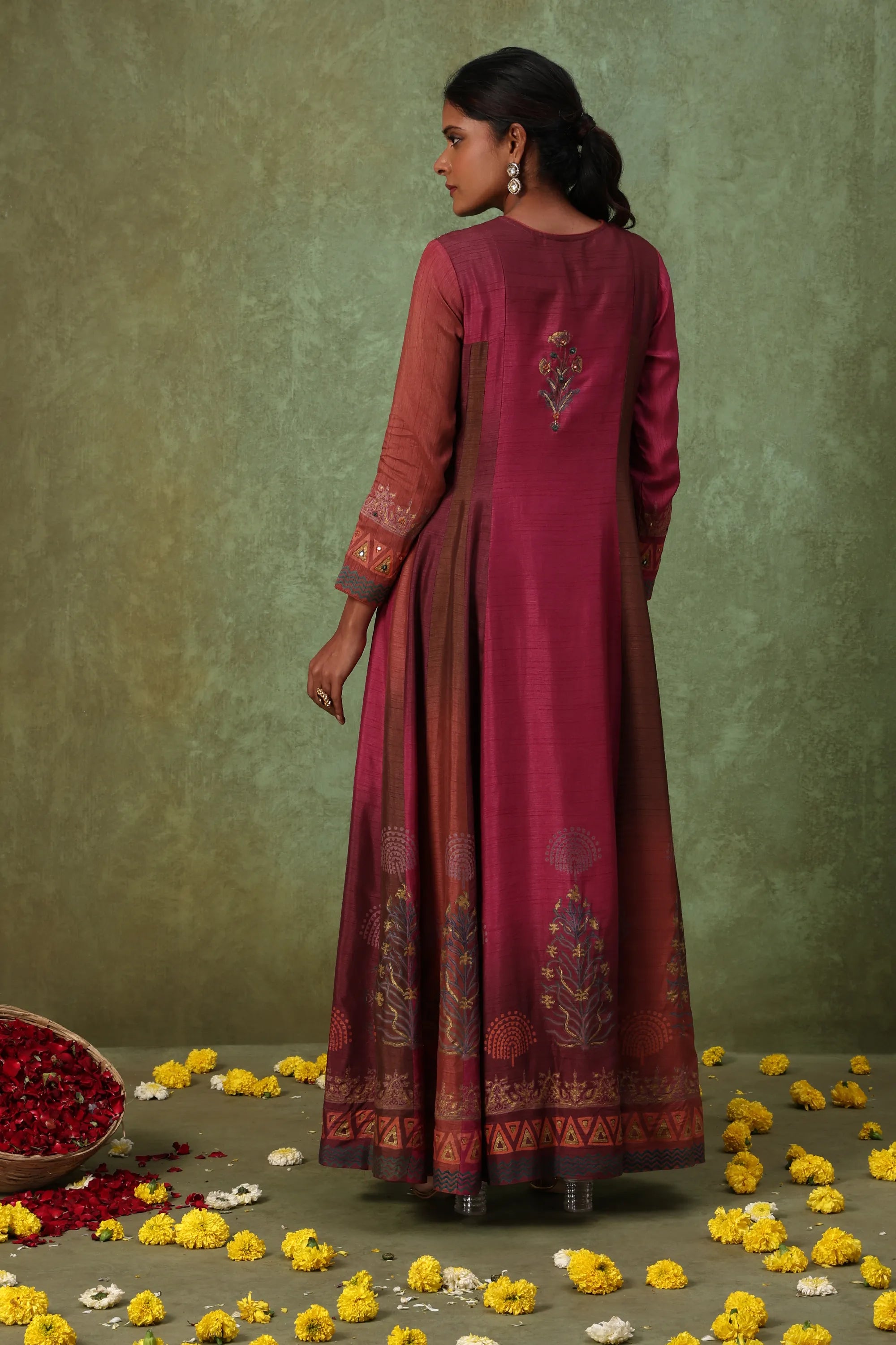 Woman in a maroon traditional outfit with floral patterns standing against a green background.