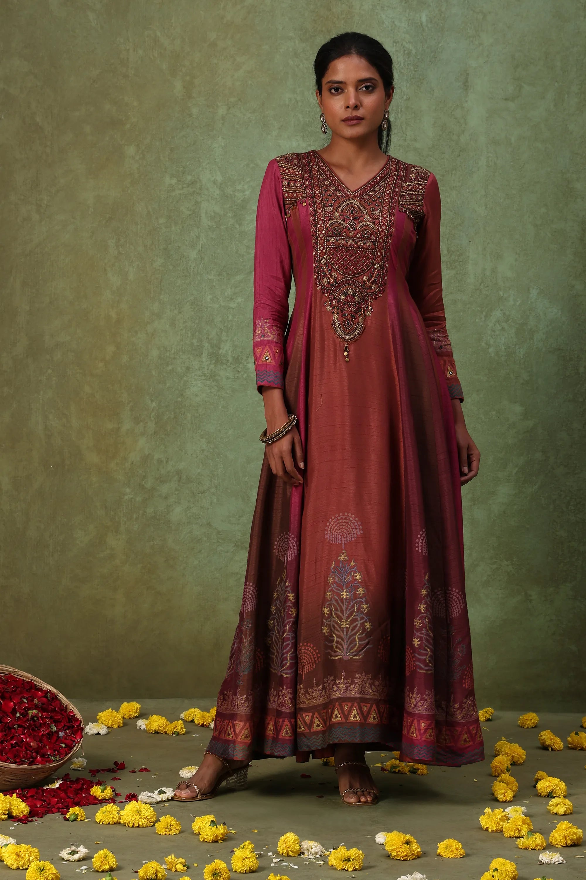 Woman wearing a long, patterned dress against a green background with flowers on the ground.