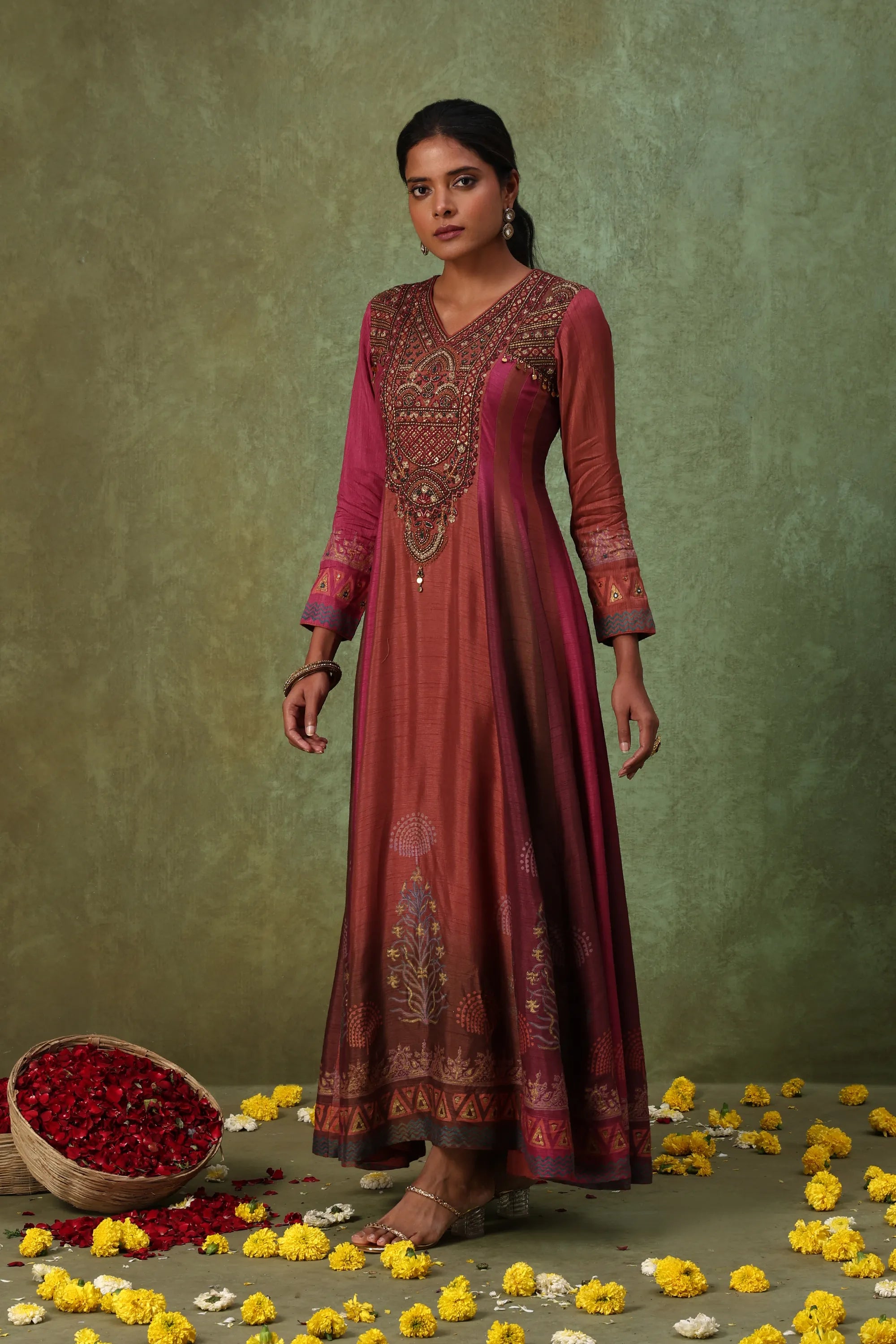 Woman wearing a long, dark red embroidered dress standing against a green background with flowers on the ground.