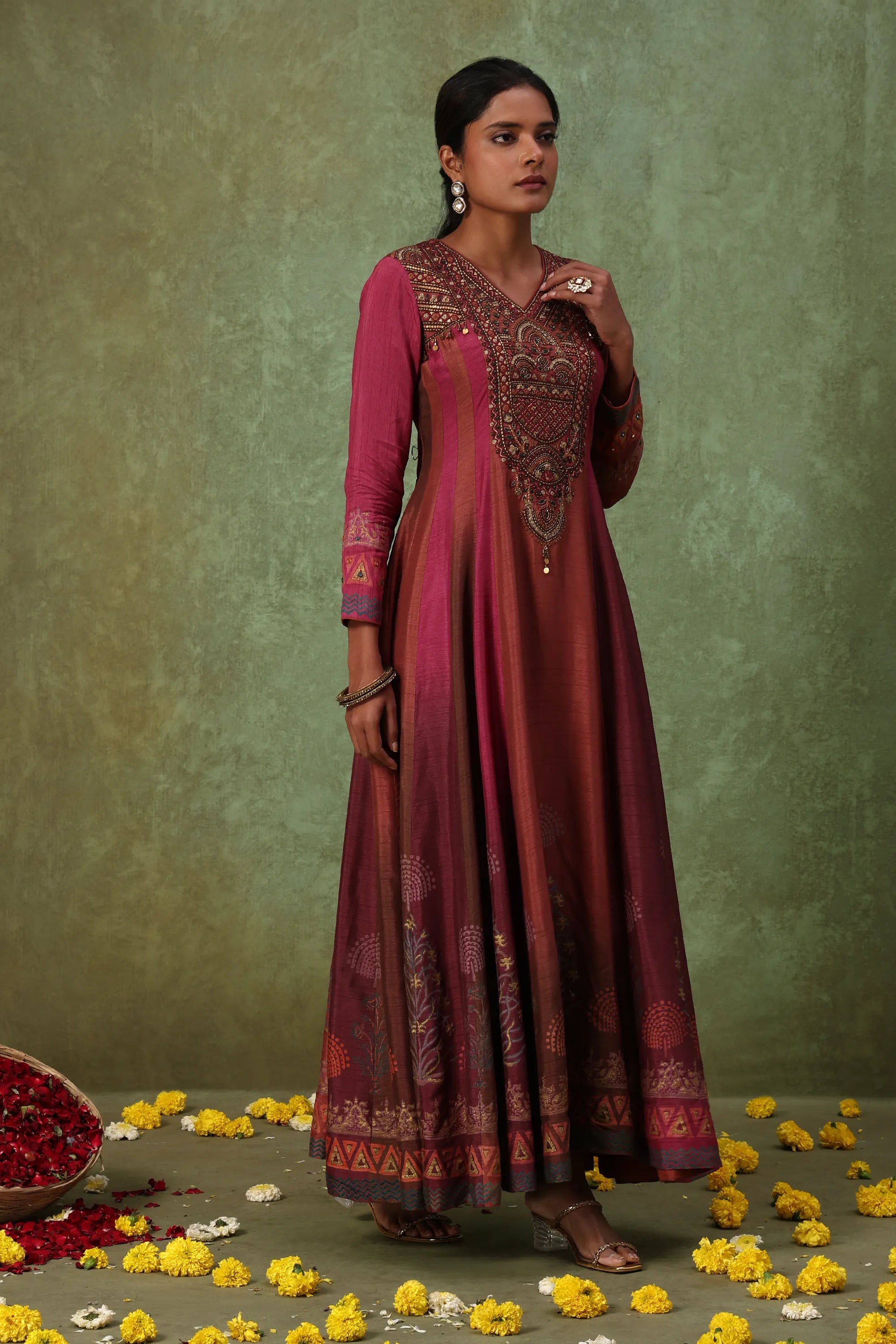 Woman in a traditional maroon and pink outfit with intricate patterns, standing against a green background.