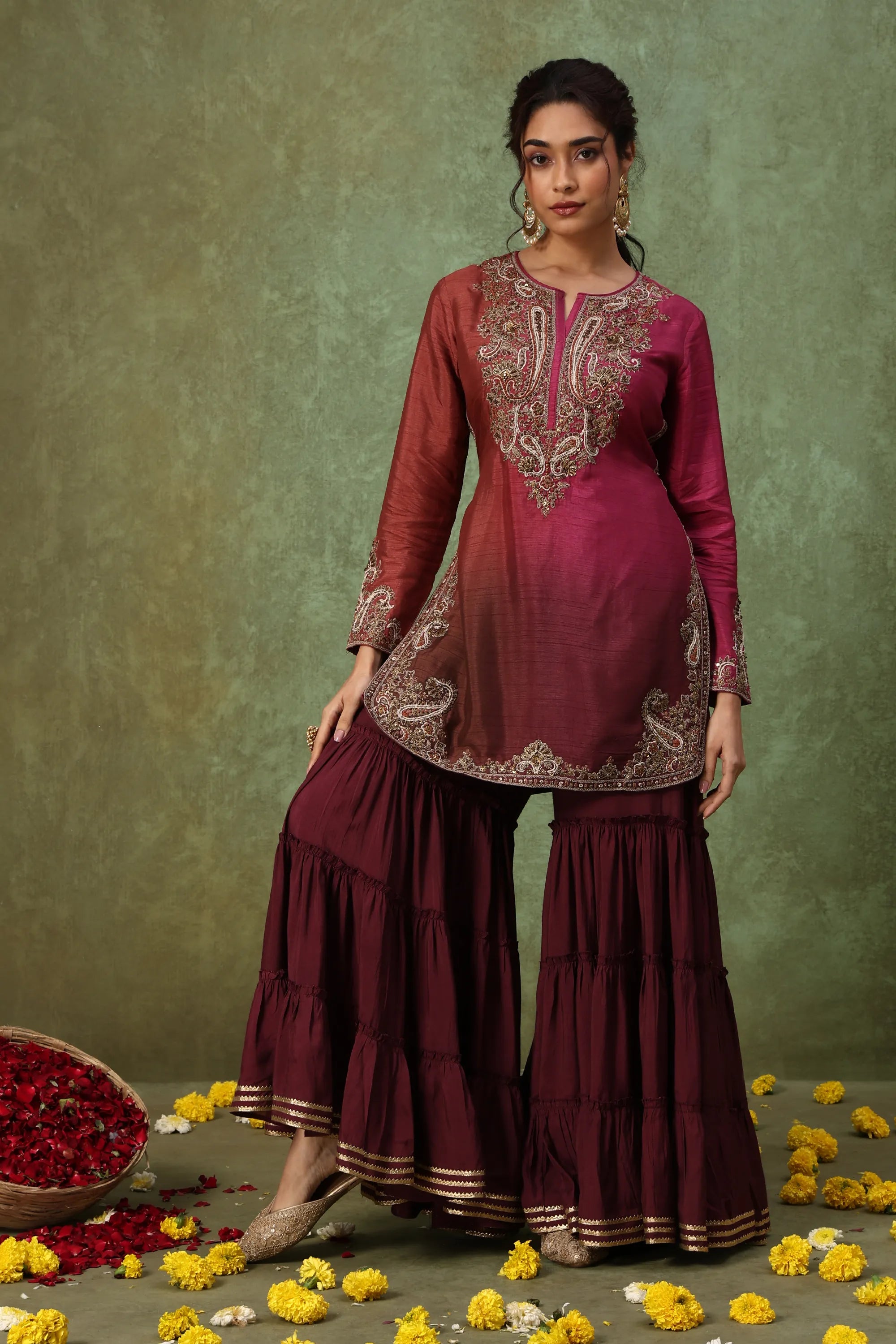 Woman wearing a maroon traditional outfit with intricate embroidery against a green background.