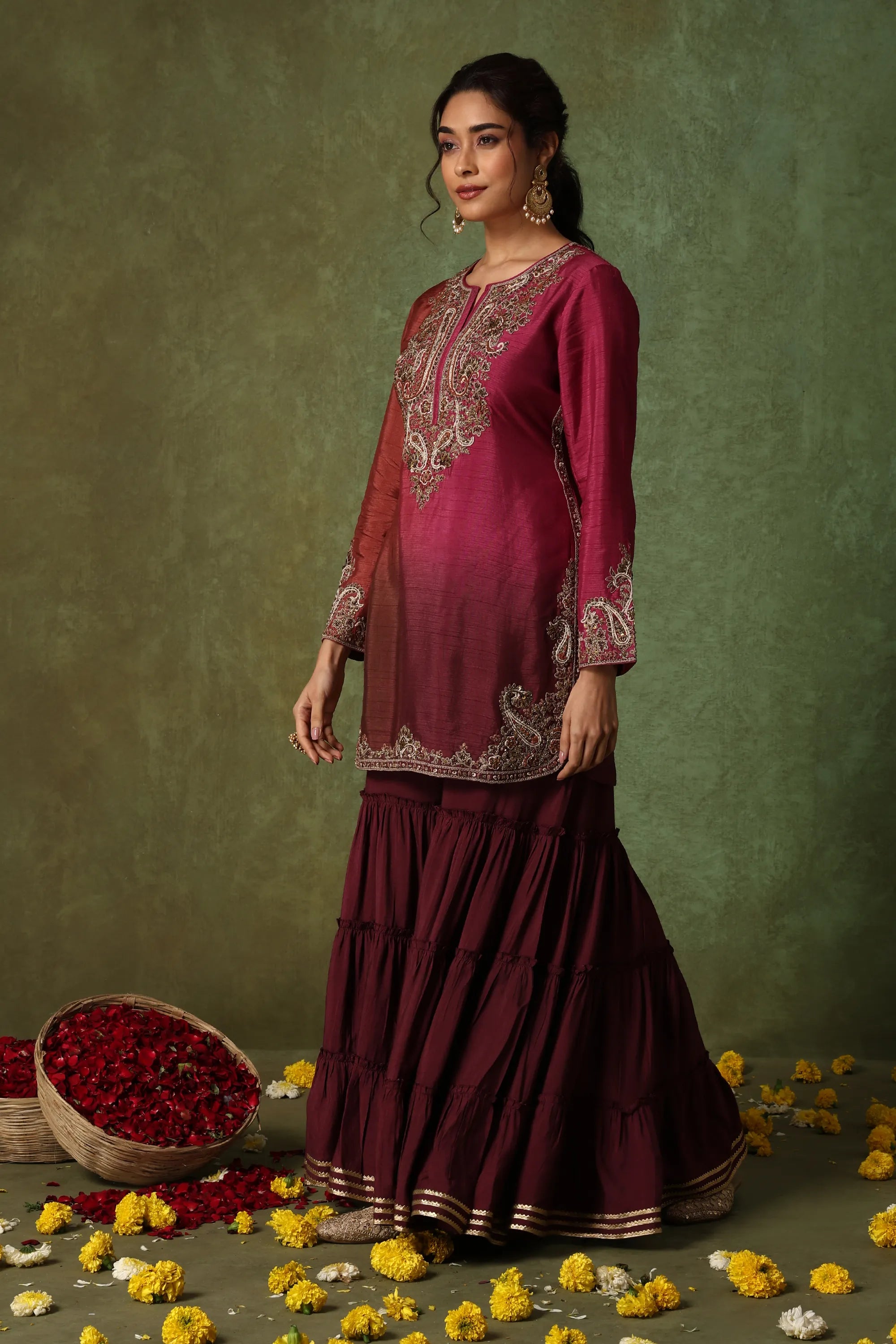 Woman in a maroon embroidered outfit standing against a green background with flowers around.