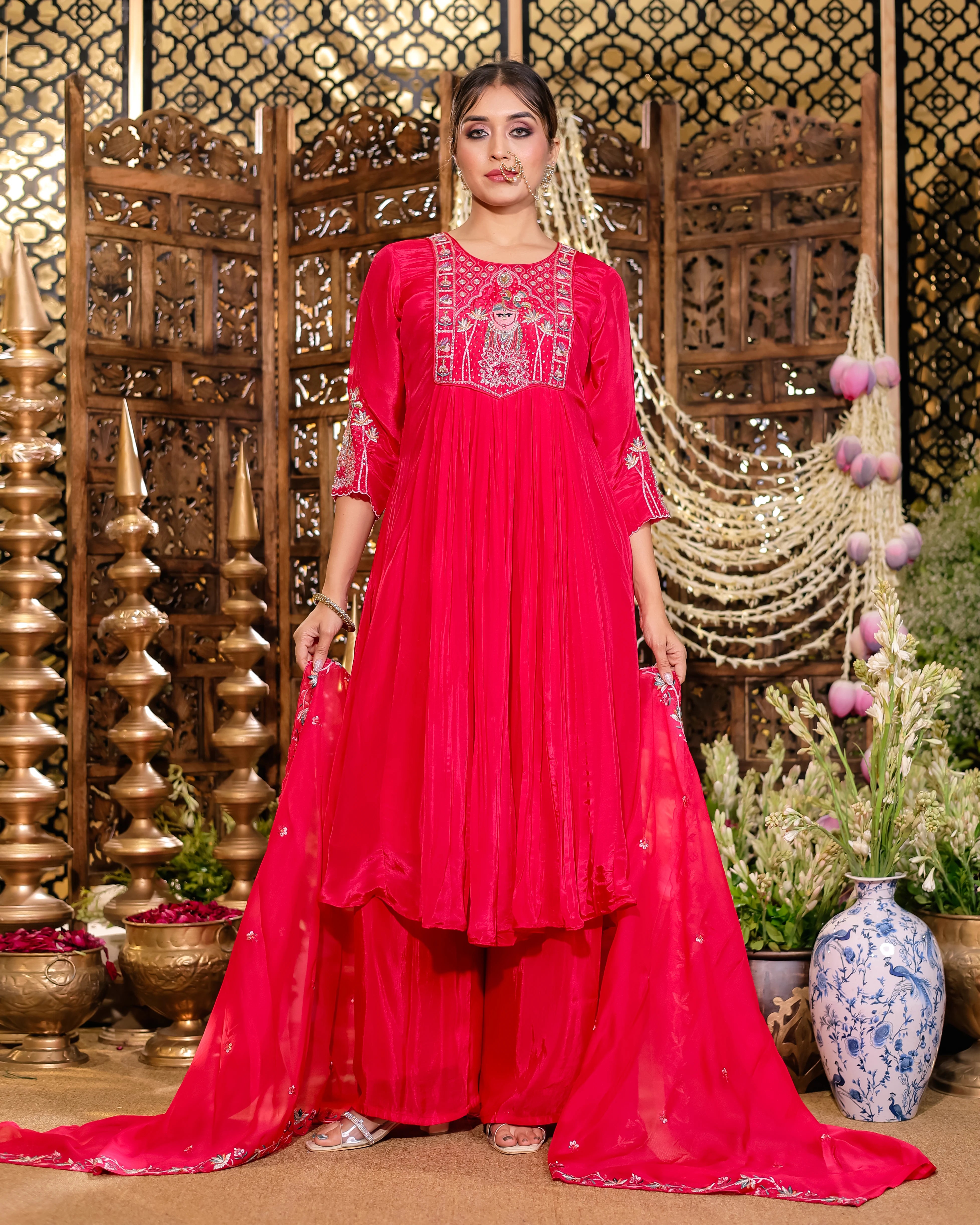 Woman in a bright pink traditional outfit standing in a decorative setting with plants and vases.