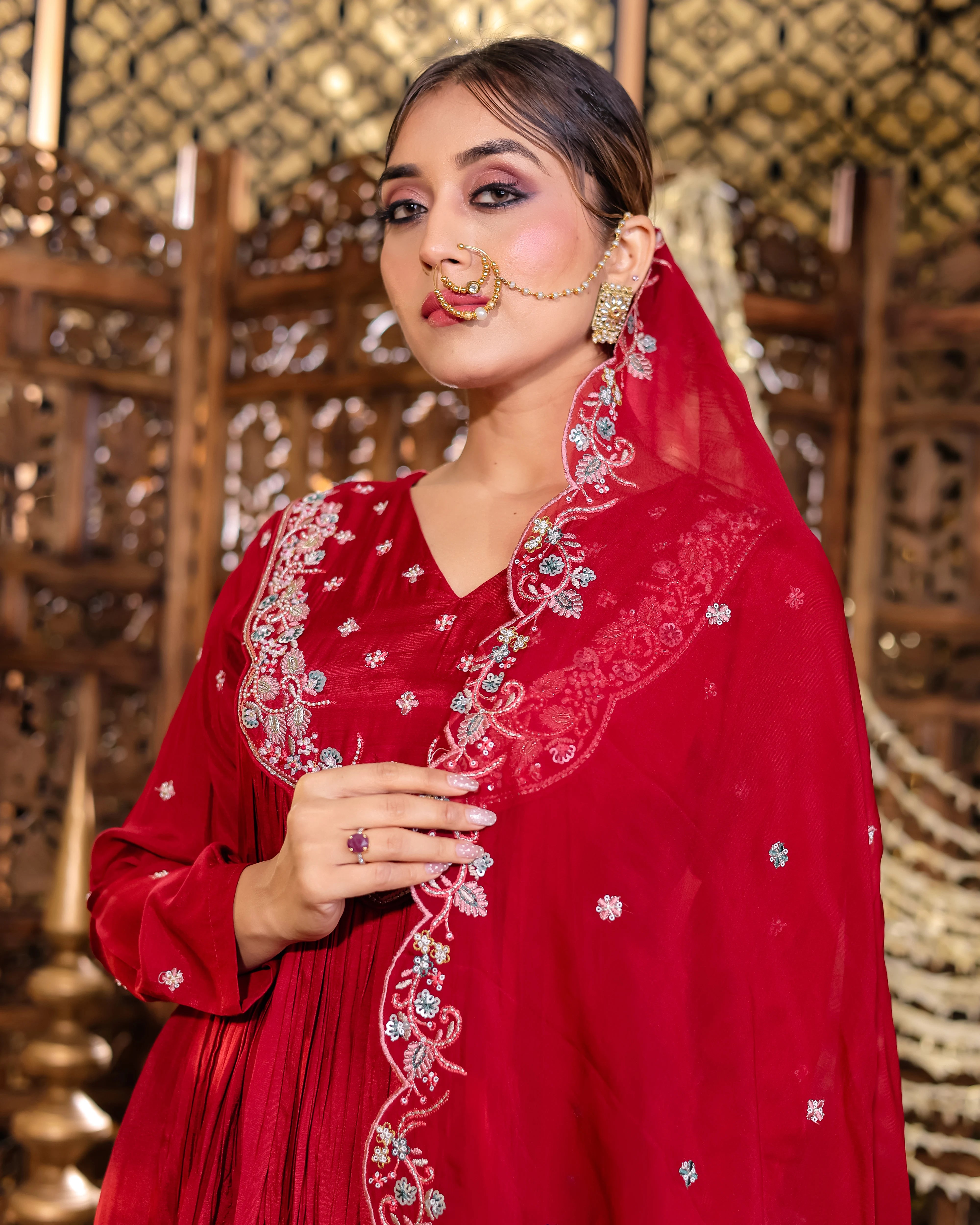 Woman wearing a red embroidered traditional outfit with a decorative necklace against a textured wall.