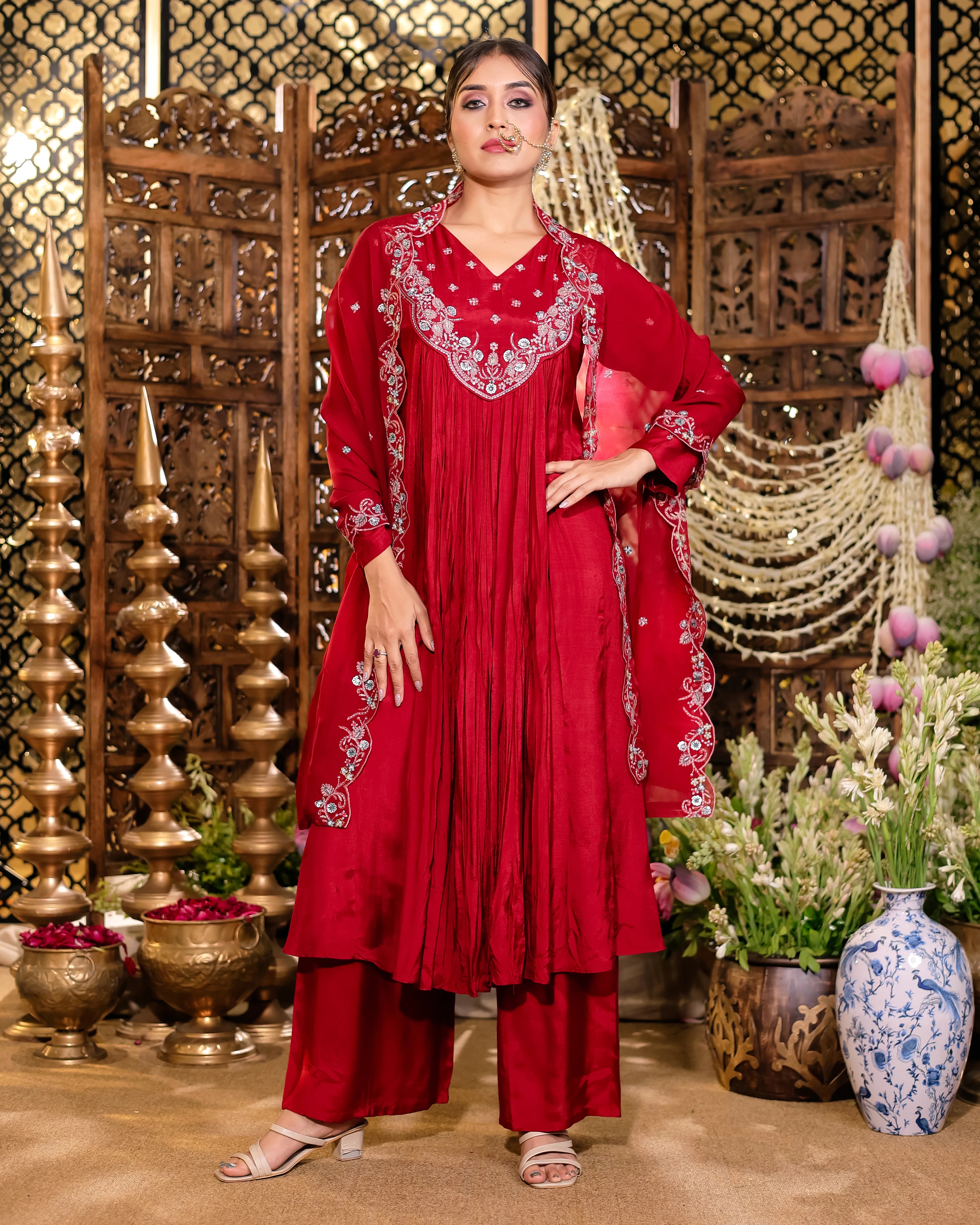 Woman in a red embroidered traditional outfit standing in a decorative setting with plants and decor.