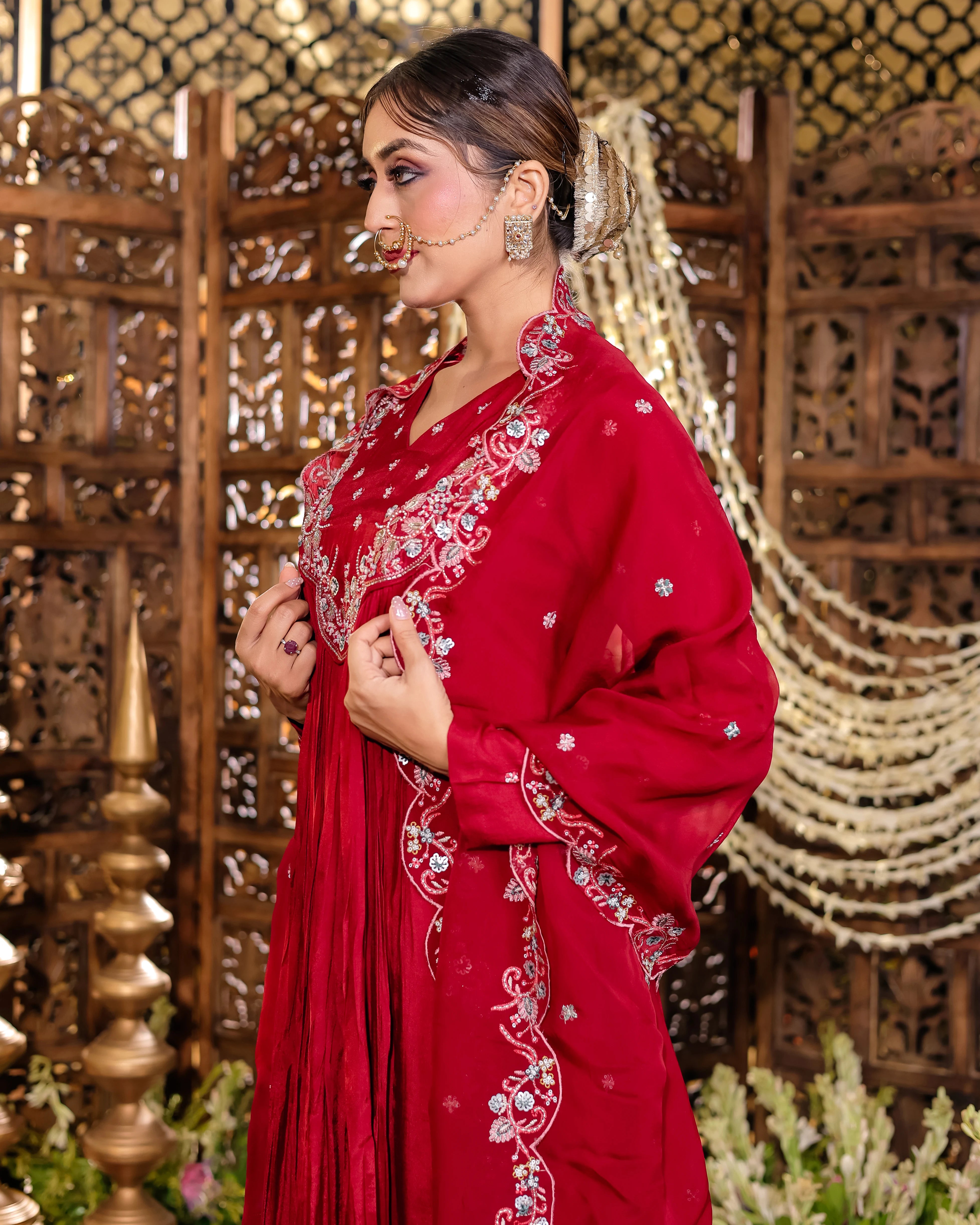 Woman in a red embroidered traditional outfit with intricate designs, standing against an ornate wooden and decorative background.