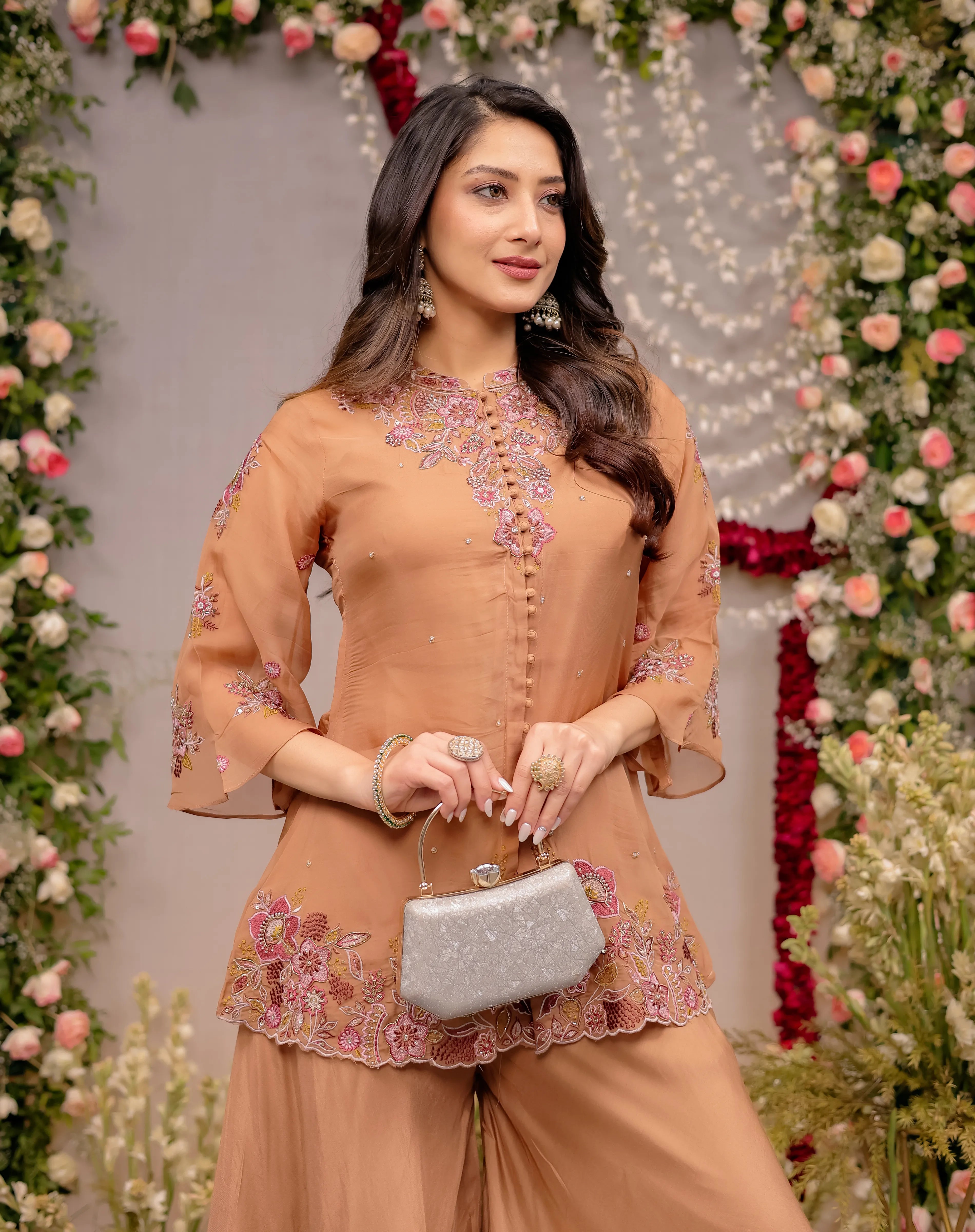 Woman in a peach traditional outfit with floral embroidery, holding a silver clutch, against a decorated floral background.