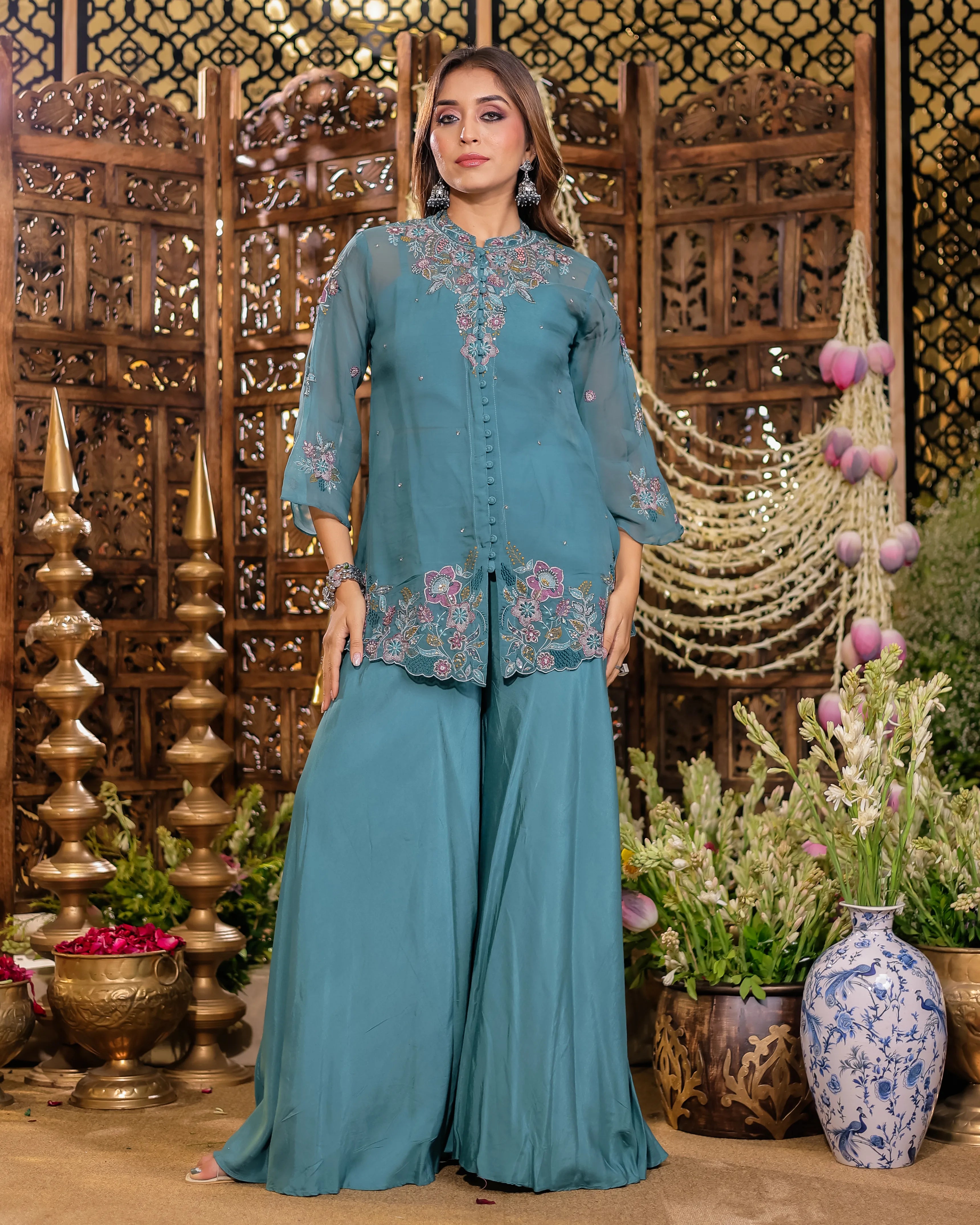 Woman in a teal traditional outfit with floral embroidery standing in front of decorative background