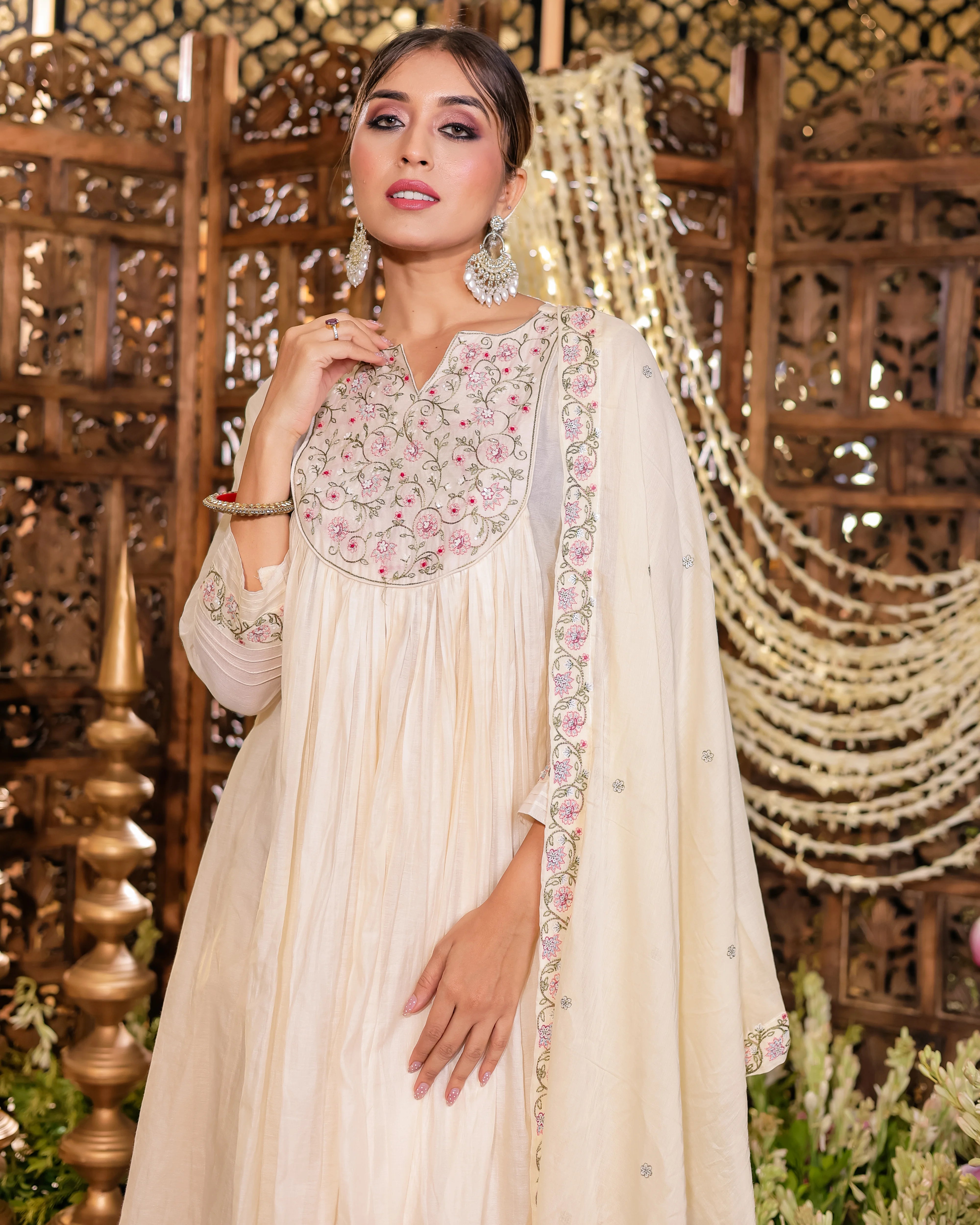 Woman in a cream-colored traditional outfit with floral embroidery against an ornate wooden backdrop.