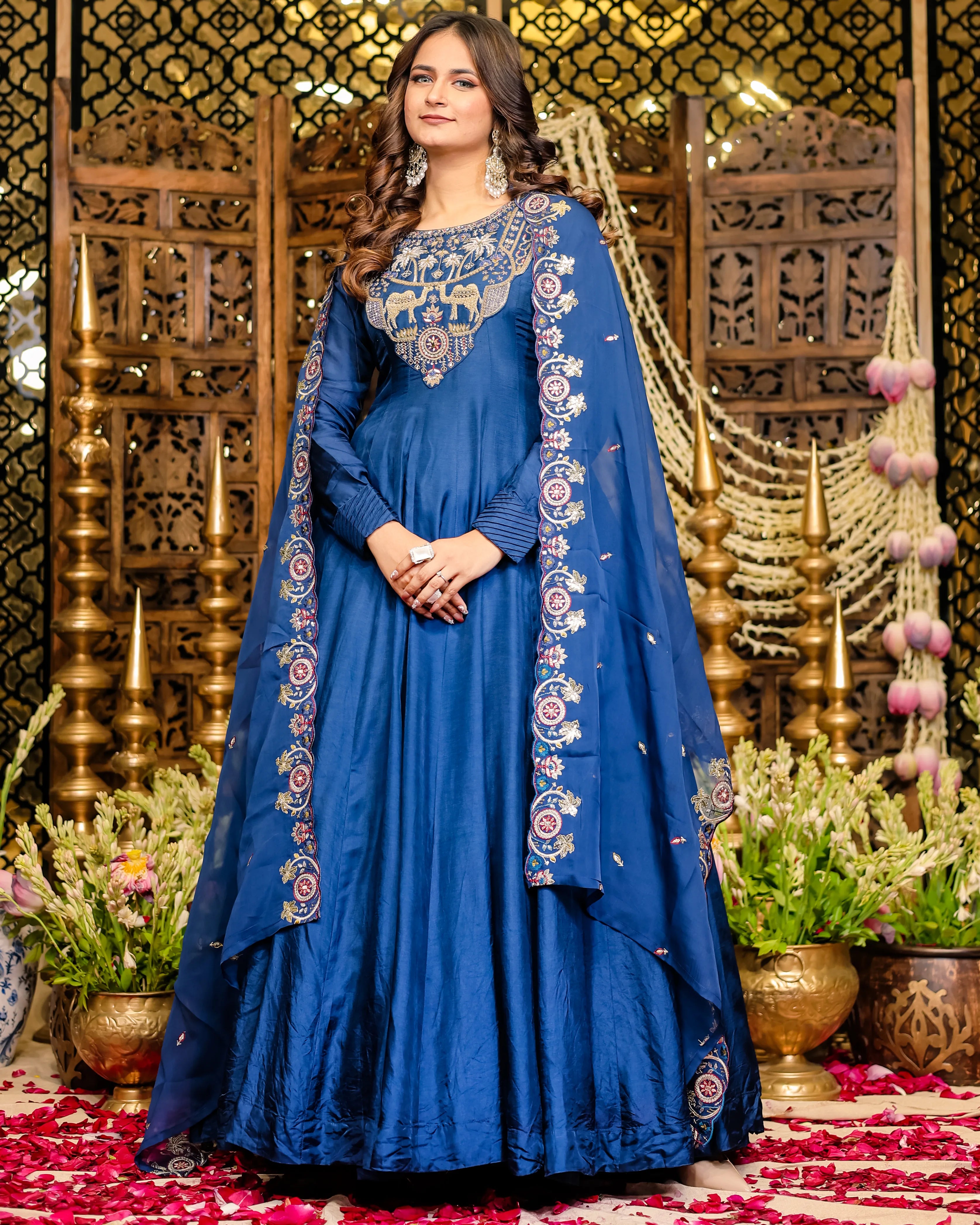 Woman in a blue embroidered traditional outfit standing in front of decorative background with plants and flowers.