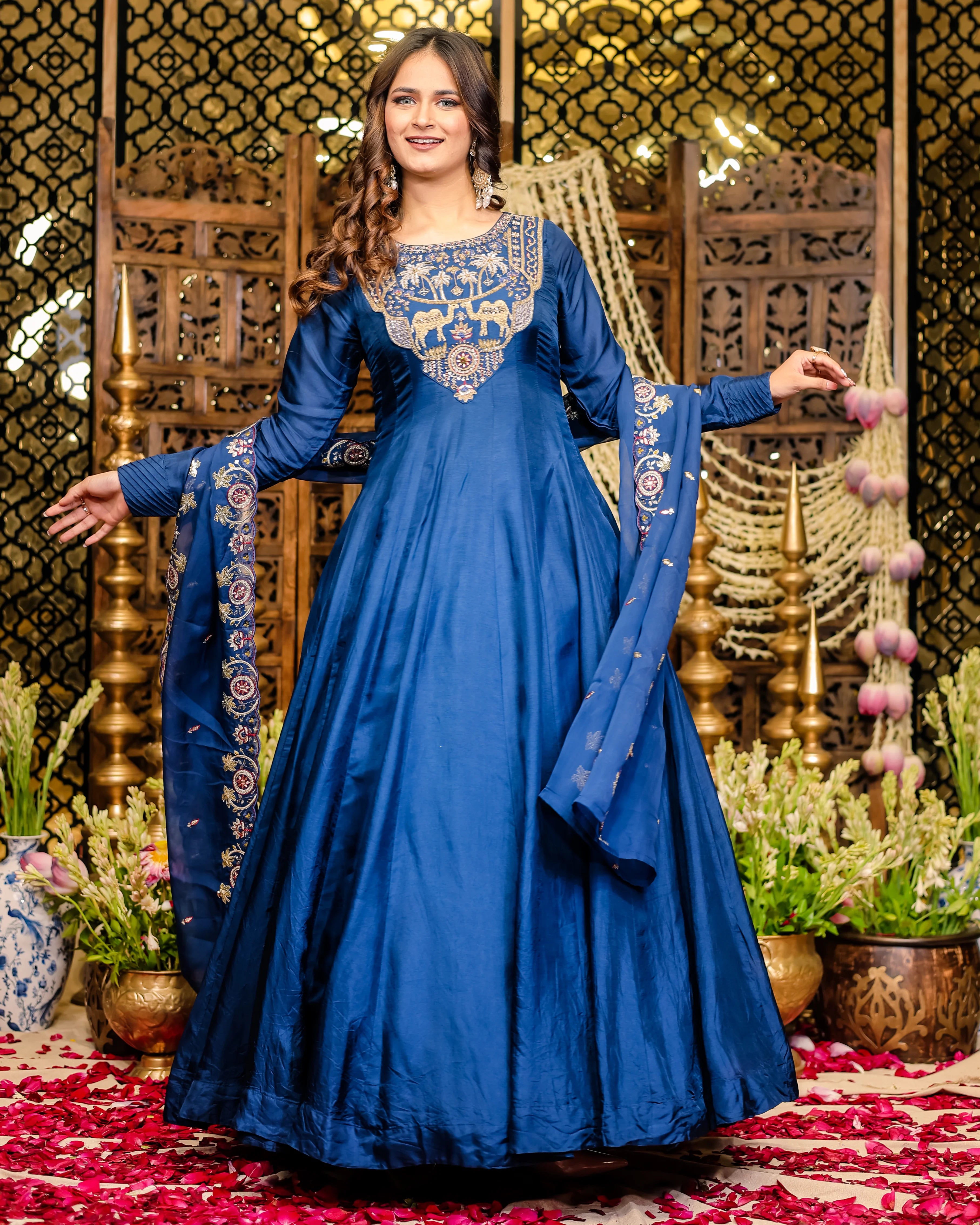 Woman in a blue embroidered traditional outfit standing in front of decorative background with plants.