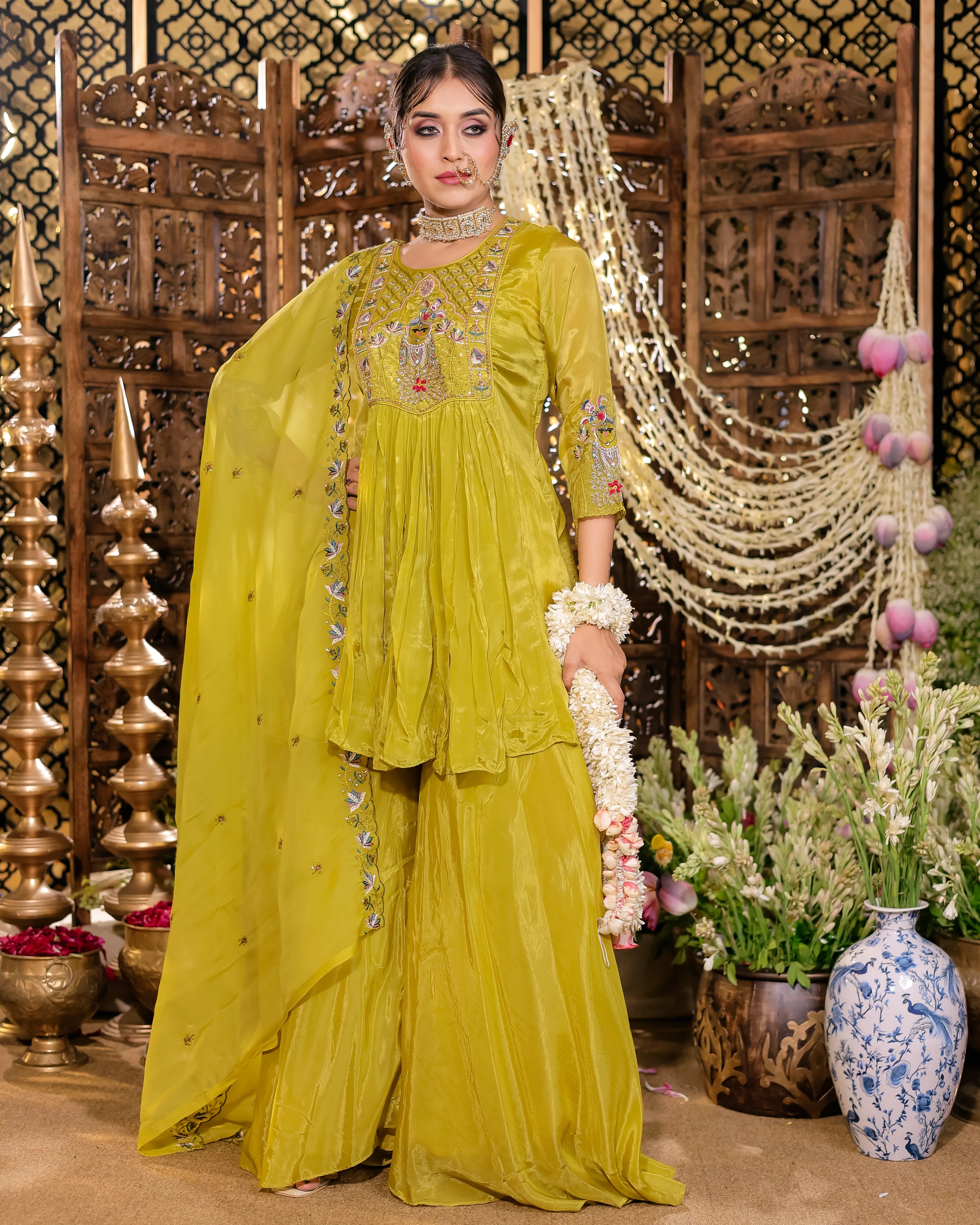 Woman in a yellow traditional outfit with intricate embroidery, standing in a decorated indoor setting.