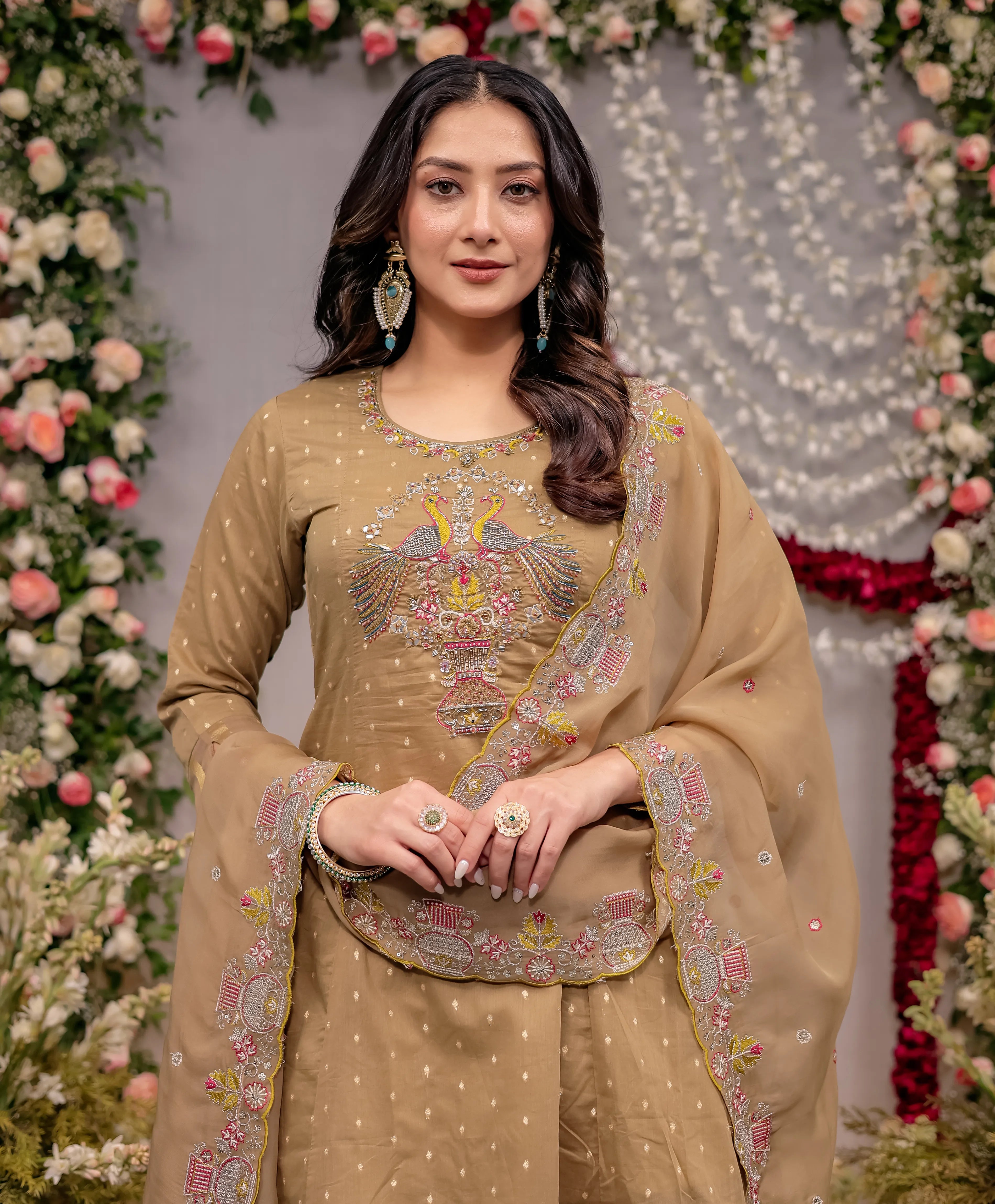 Woman in a beige embroidered traditional outfit standing in front of floral decorations.