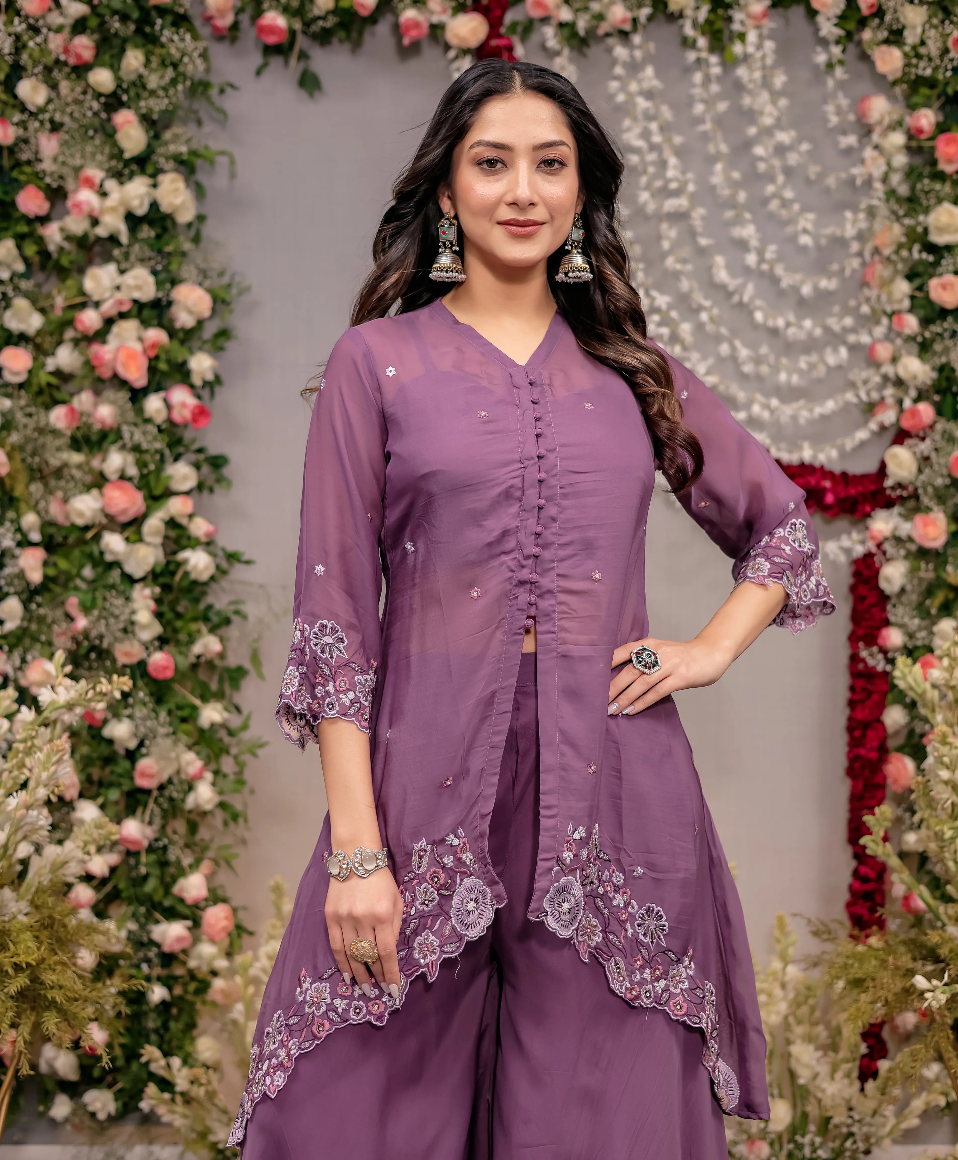 Woman in a purple traditional outfit with floral decorations against a decorated wall.