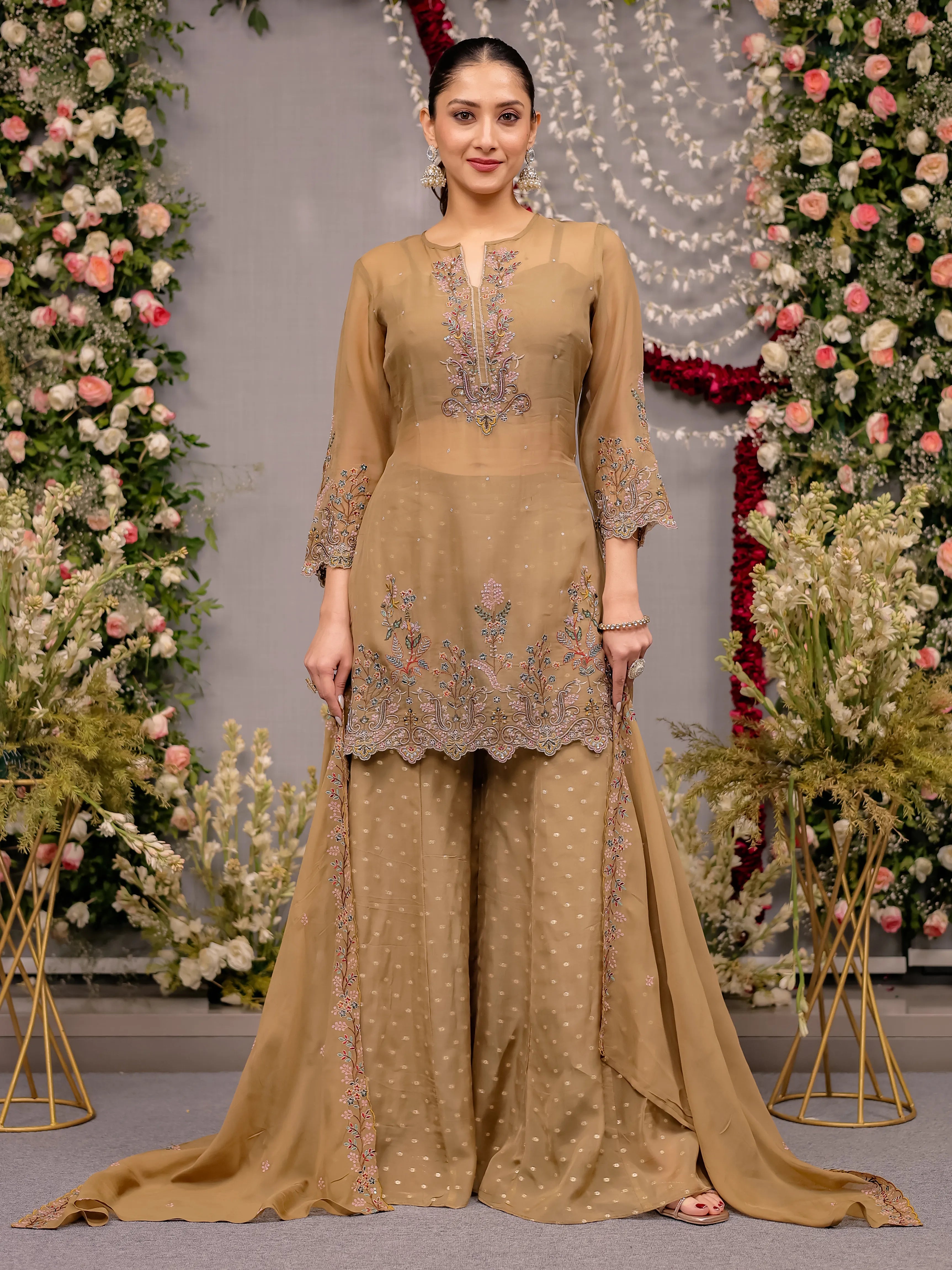 Woman in a beige embroidered traditional outfit standing in front of floral decorations.