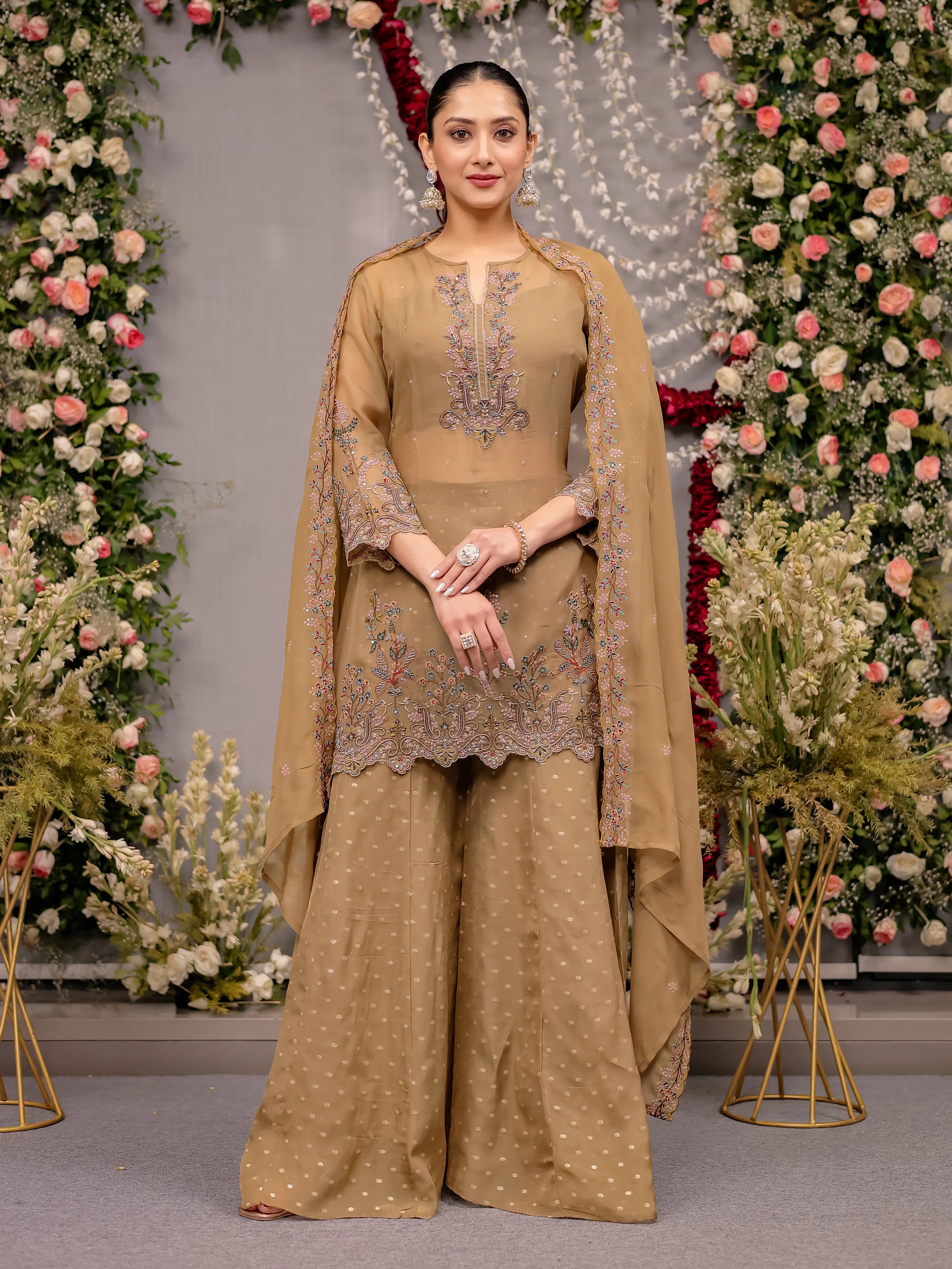Woman in a beige embroidered traditional outfit standing in front of floral decorations.