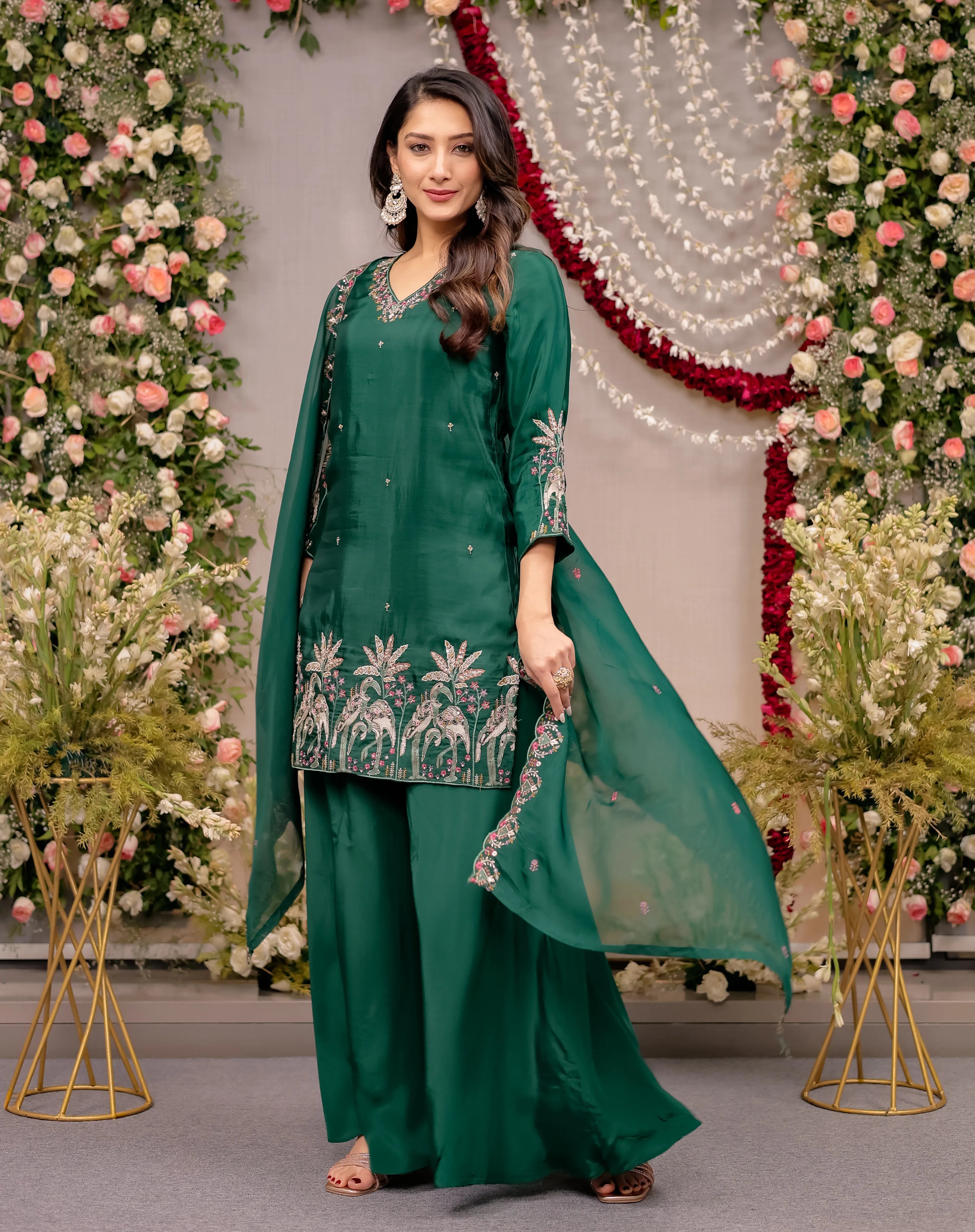 Woman in a green traditional outfit with floral embroidery against a decorated background with flowers and lights.