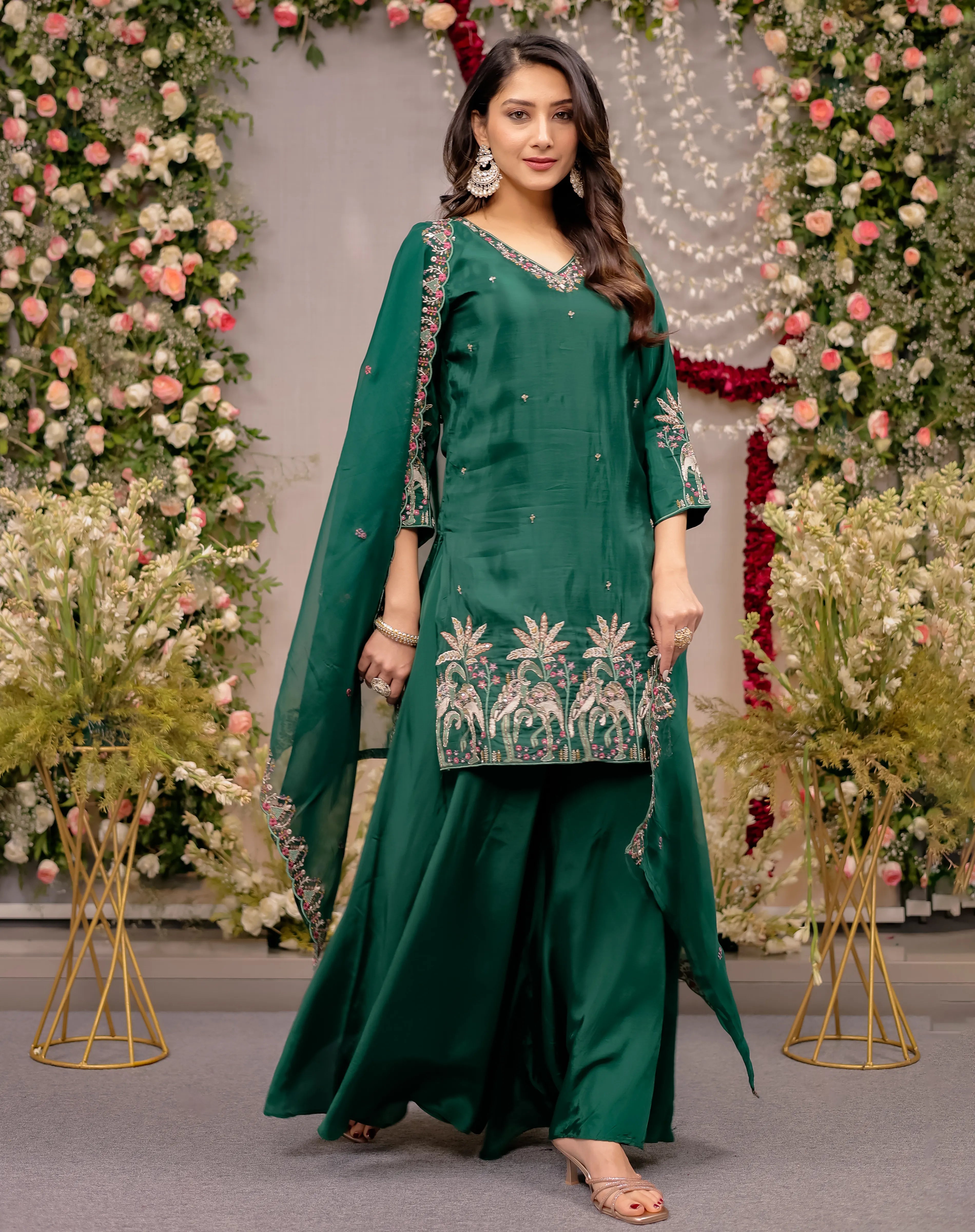 Woman in a green traditional outfit with floral embroidery standing in front of floral decorations.