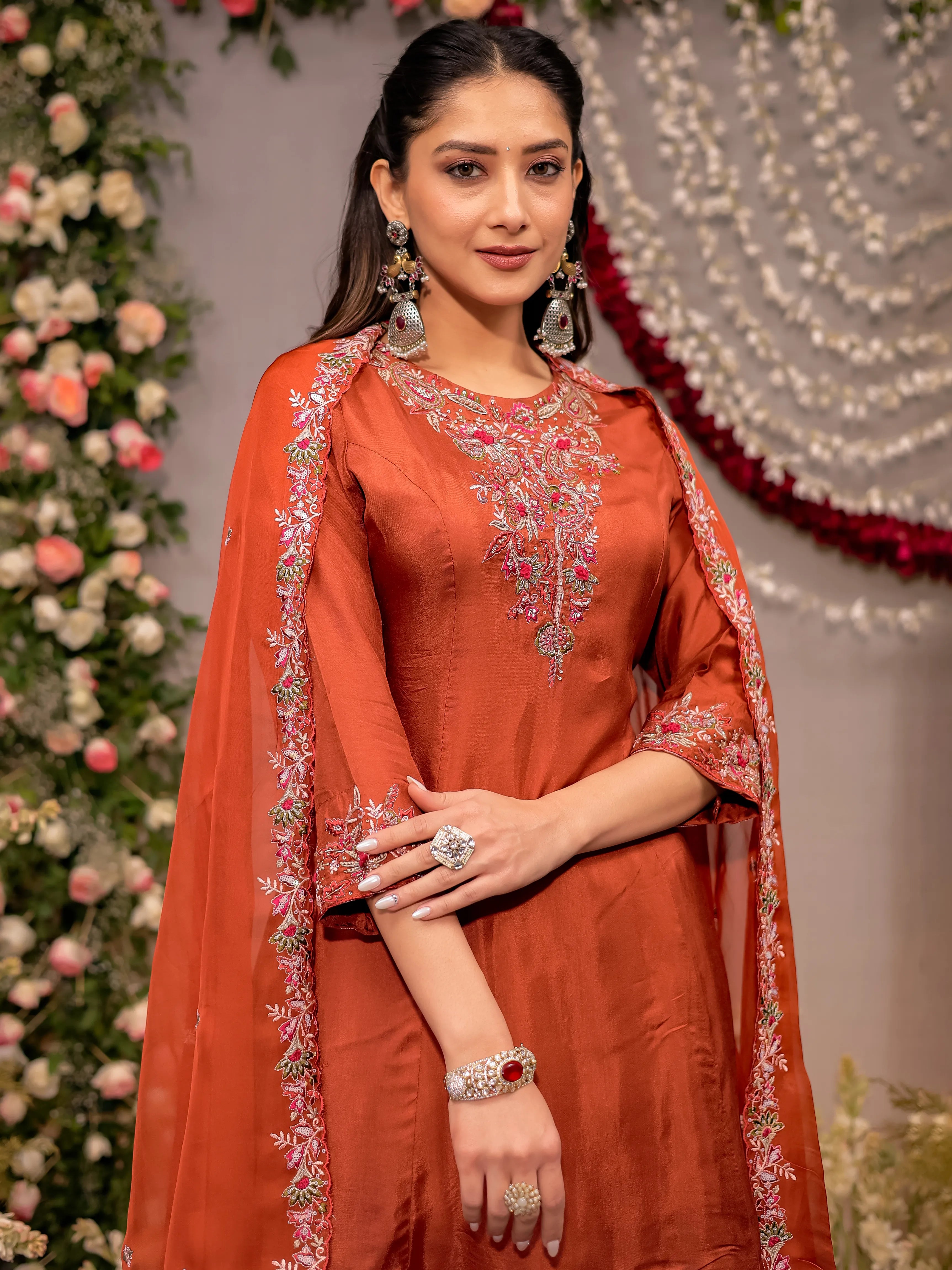 Woman in an orange traditional outfit with embroidery, standing against a decorated backdrop.