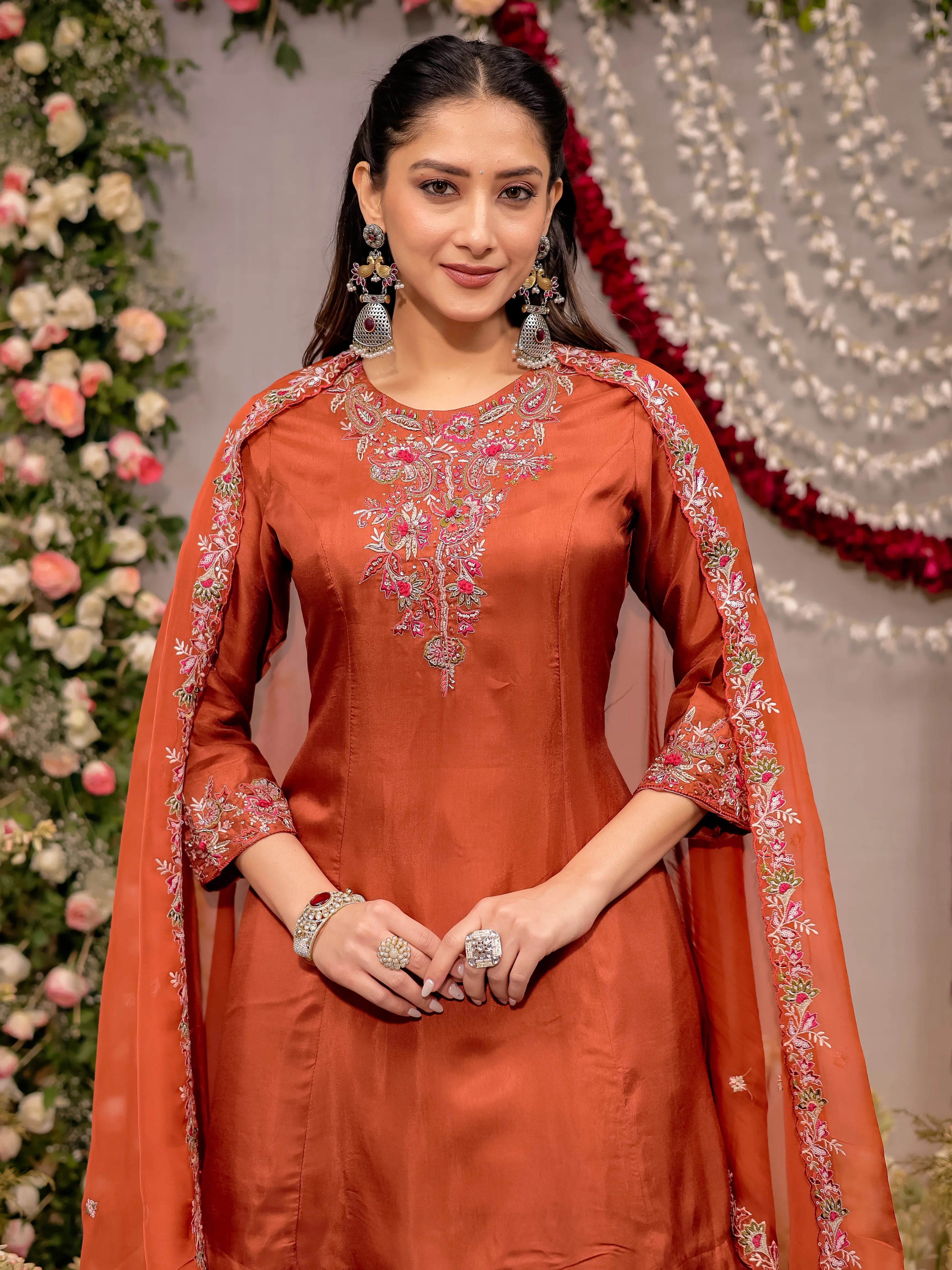 Woman in an orange embroidered traditional outfit standing against a decorated background with flowers and lights.