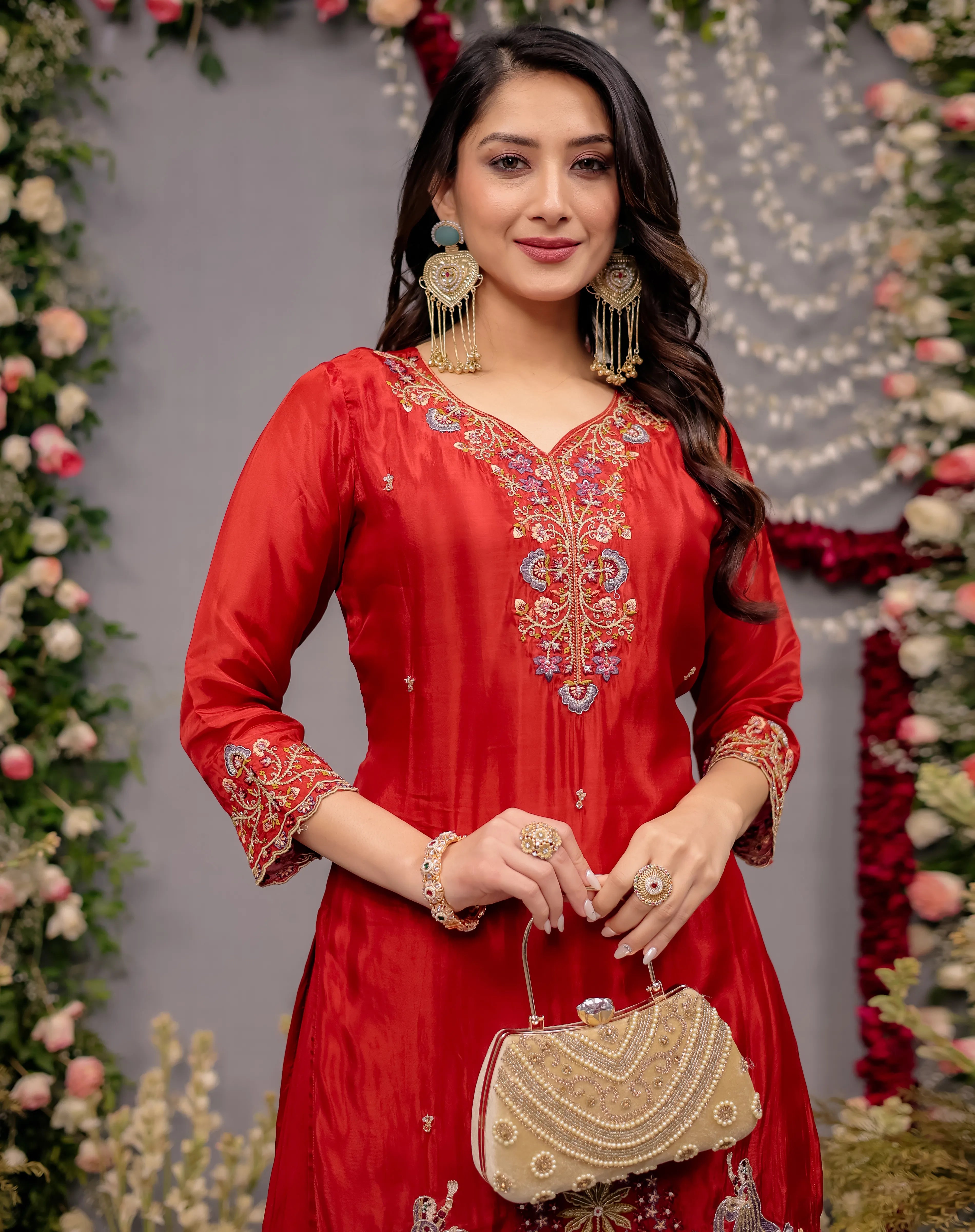 Woman in a red embroidered outfit holding a gold clutch against a floral decorated background