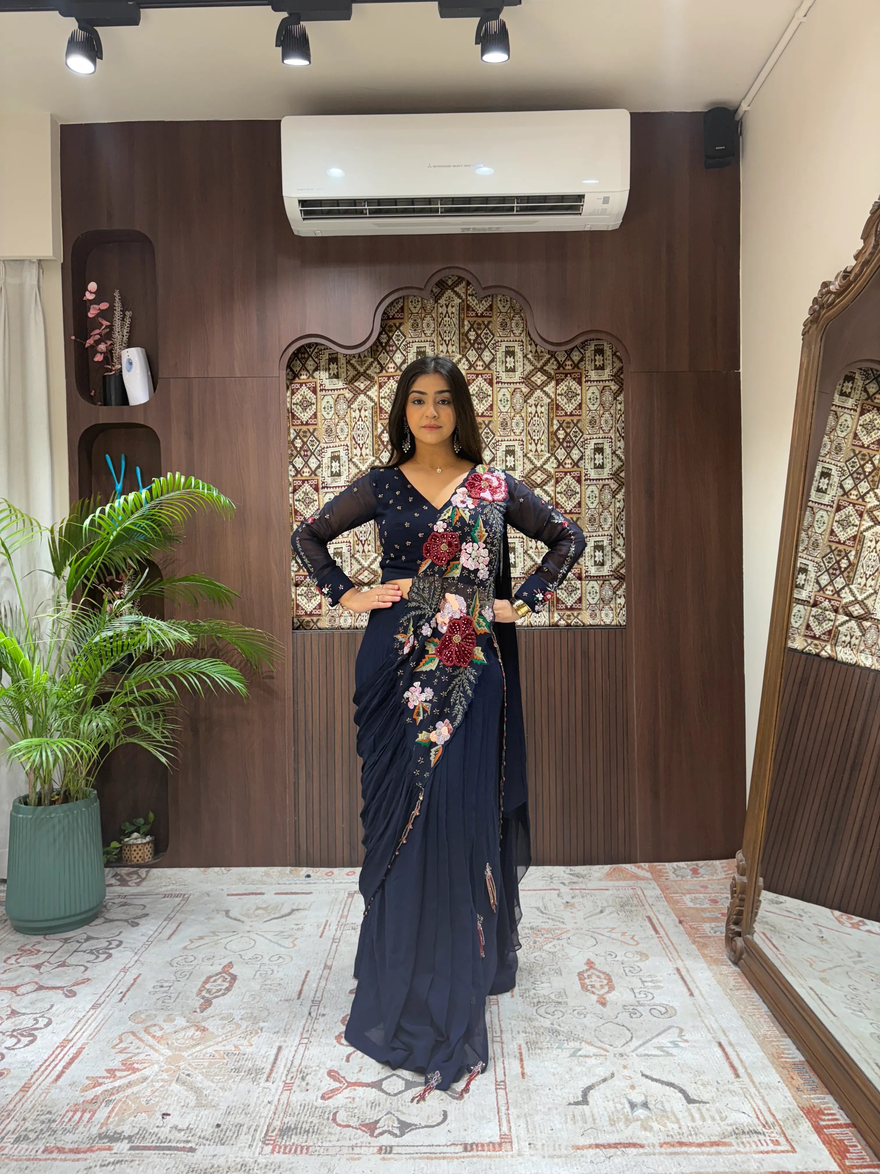 Woman in a floral saree standing in a room with decorative walls and plants.