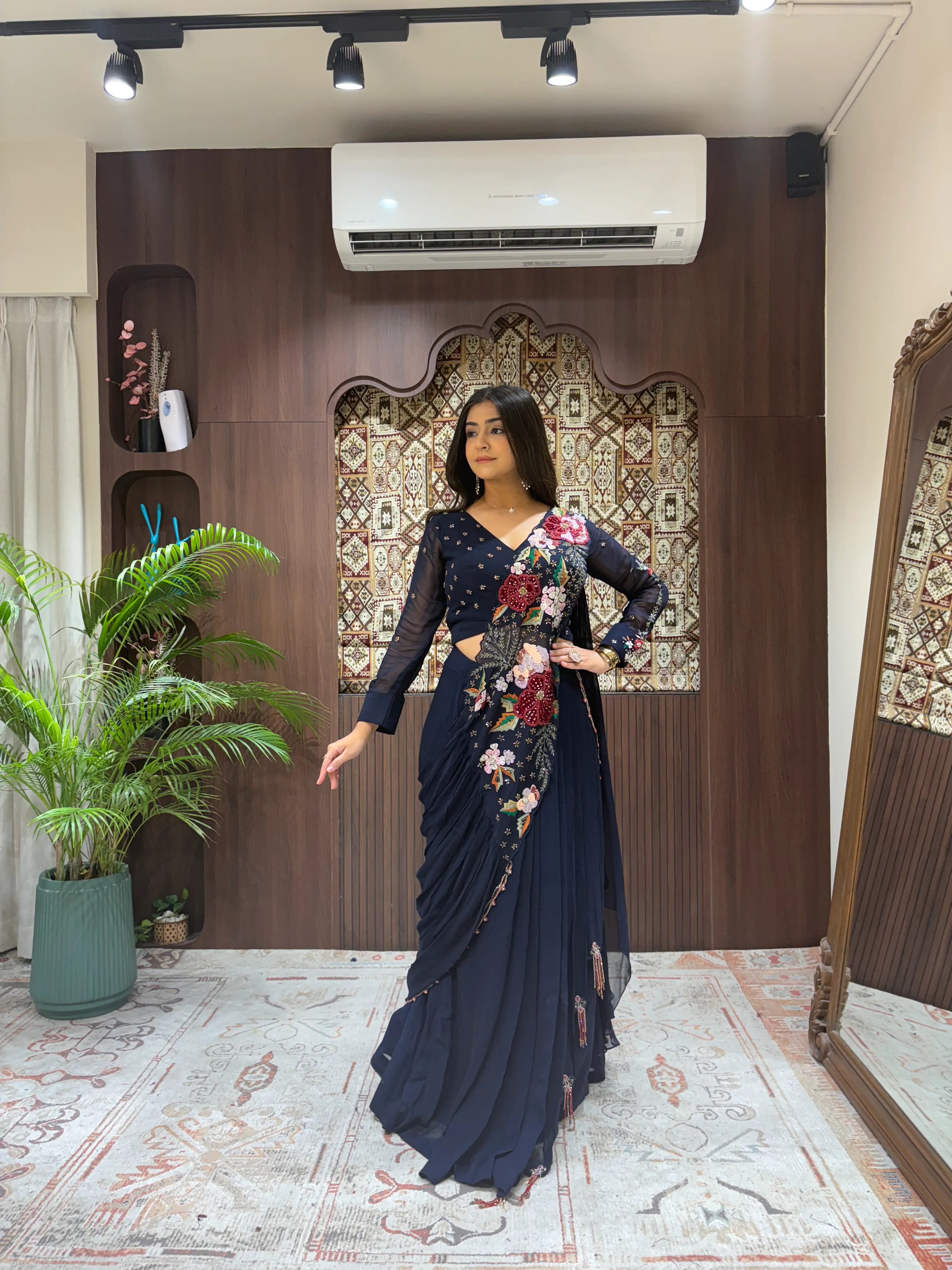 Woman in a floral saree standing in a decorated room with plants and an air conditioner.