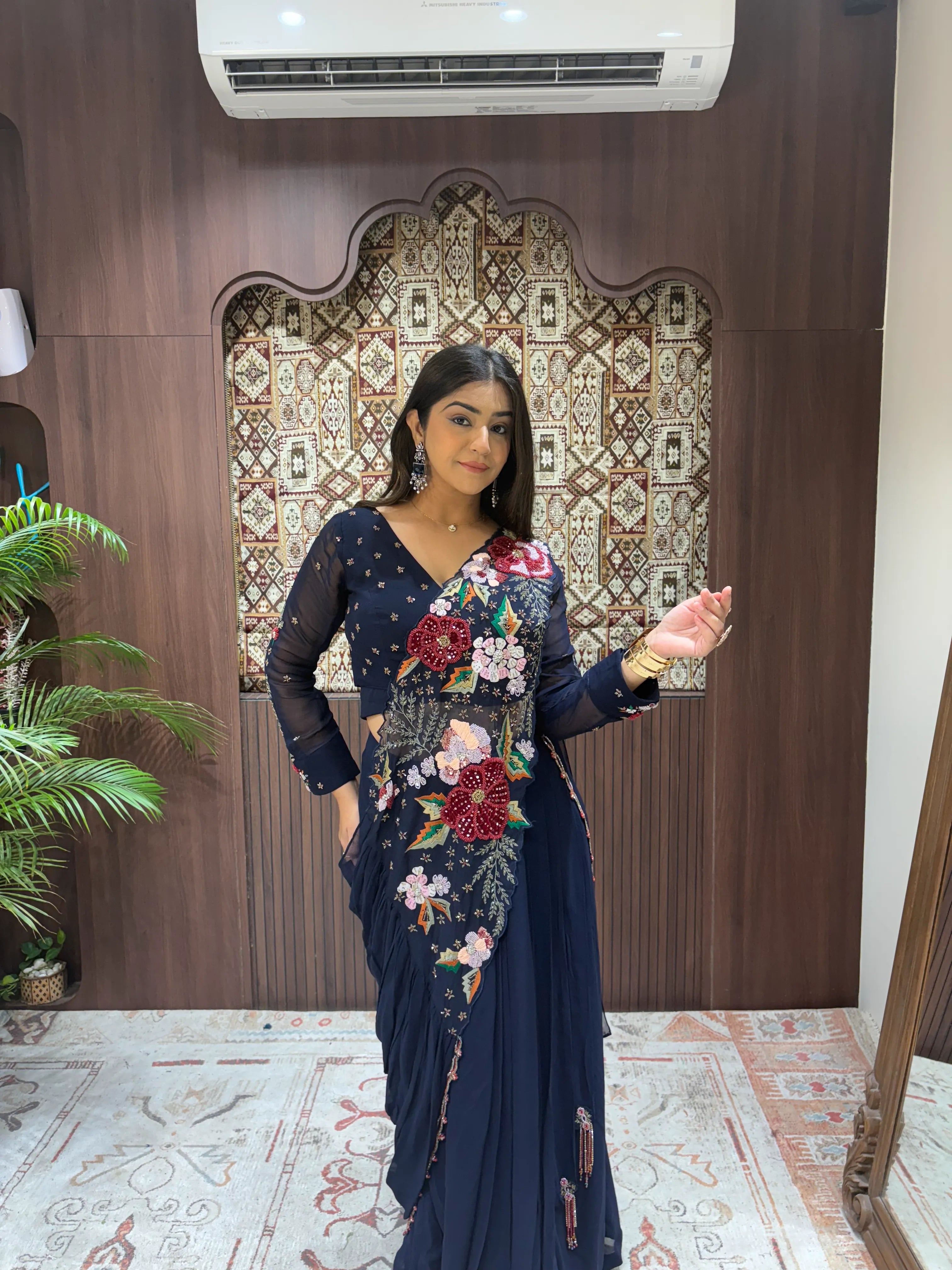 Woman in a floral saree standing in a room with decorative wall and plant.